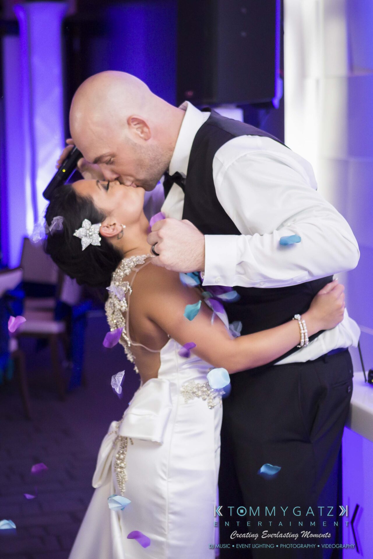 135 - TOMMY GATZ ENTERTAINMENT A couple shares a kiss at a wedding, surrounded by falling confetti. The bride wears a white gown and the groom is in a vest and tie.