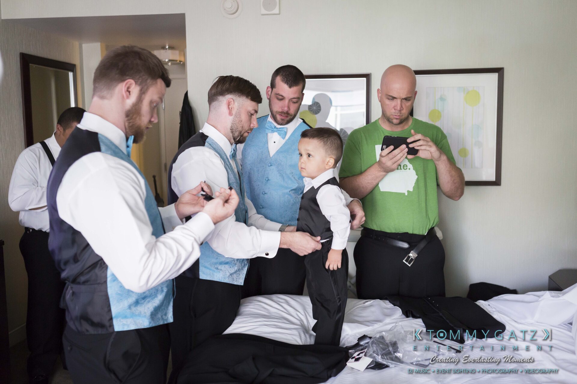 171 - TOMMY GATZ ENTERTAINMENT A group of men and a small boy in formal wear prepare for an event in a room. One man adjusts the boy's suit, while another uses a phone.