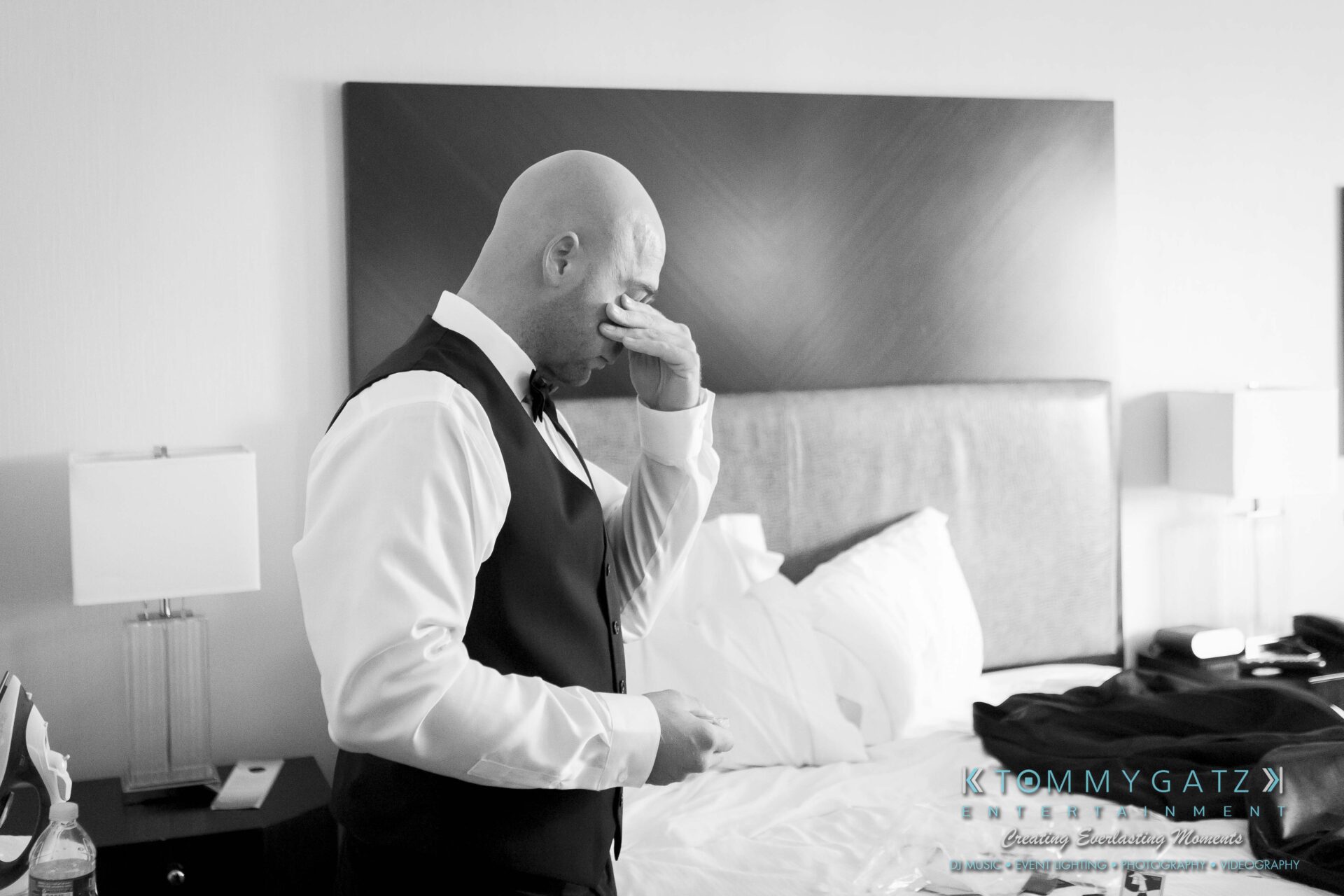 191 - TOMMY GATZ ENTERTAINMENT A man in formal attire wipes his eye in a bedroom, with an unmade bed in the background and a nightstand on the left.