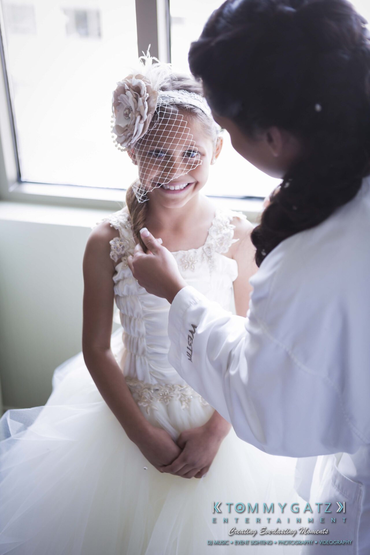 211 - TOMMY GATZ ENTERTAINMENT Bride adjusts young girl's dress in a sunlit room. Girl wears a white dress with floral embellishments and a netted headpiece.