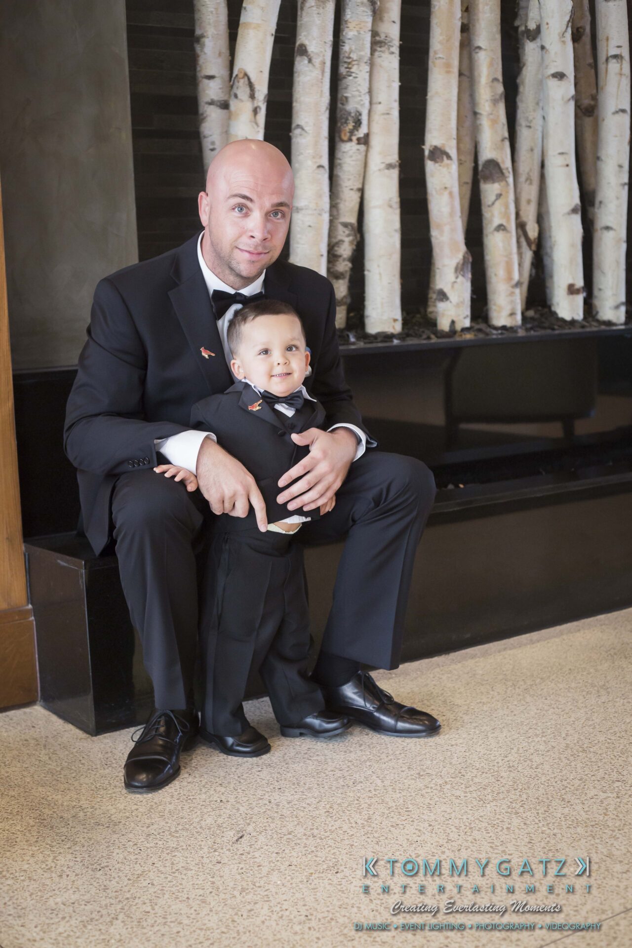 231 - TOMMY GATZ ENTERTAINMENT A man and a child in matching suits pose together in front of a birch bark wall.