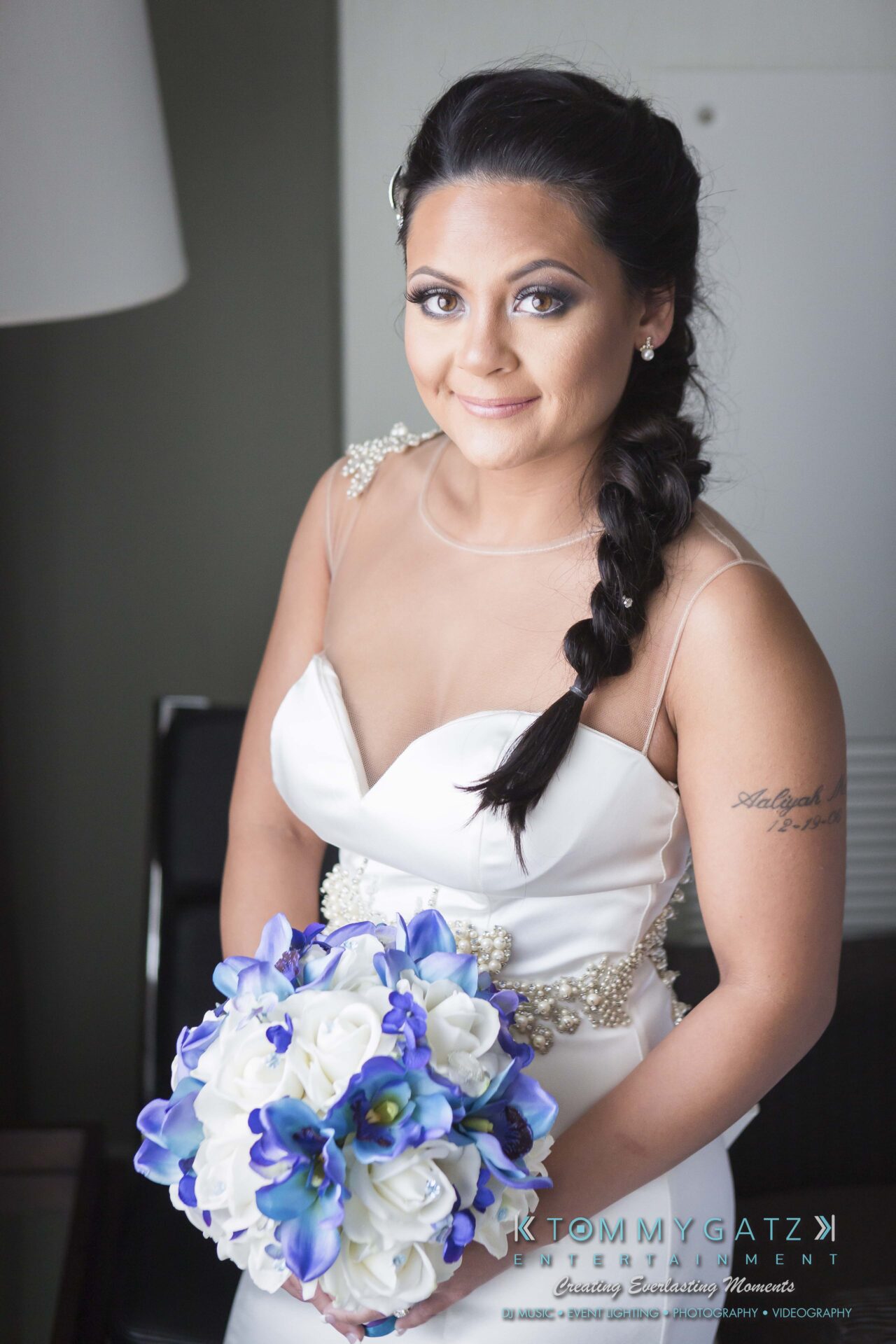 261 - TOMMY GATZ ENTERTAINMENT A woman in a white wedding dress holds a bouquet of blue and white flowers, standing indoors.