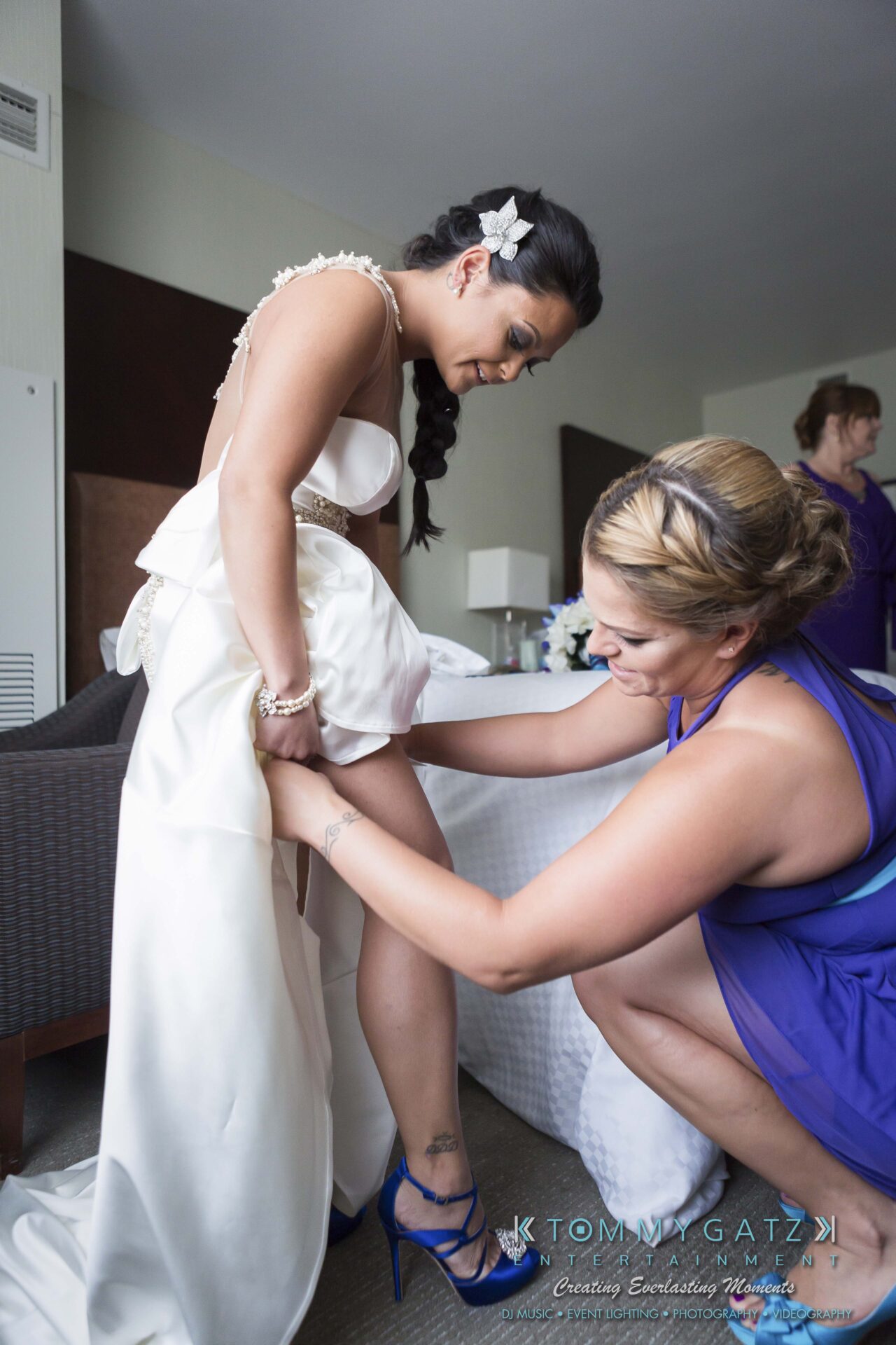 271 - TOMMY GATZ ENTERTAINMENT A woman in a white dress is helped by another woman in a blue dress, adjusting her outfit. They are in a room with a sofa and a person in the background.