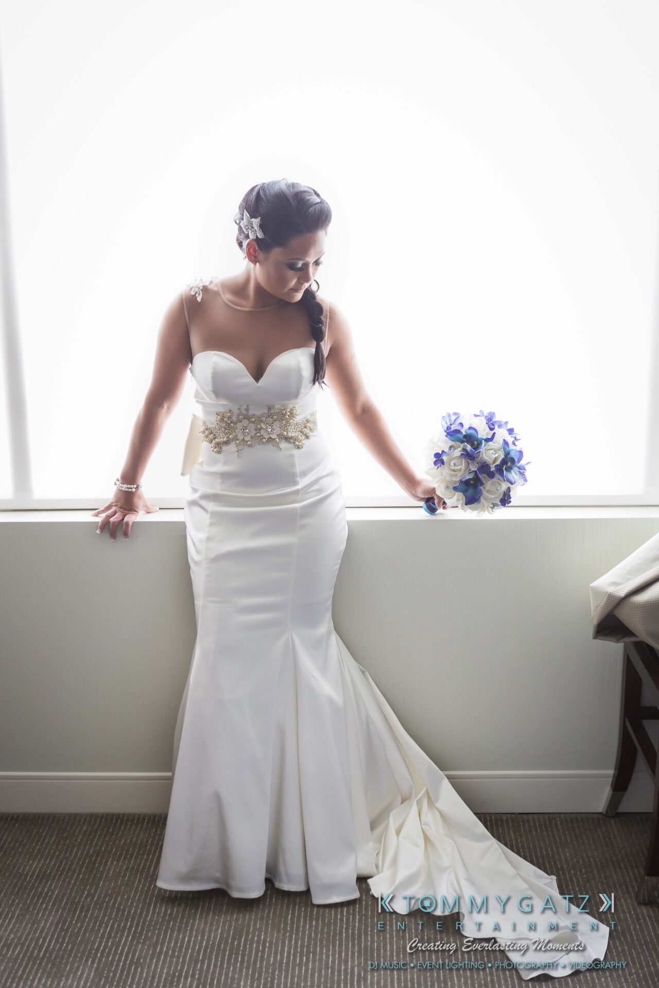 281 - TOMMY GATZ ENTERTAINMENT A bride in a white wedding gown holds a bouquet of blue and white flowers, standing by a bright window.