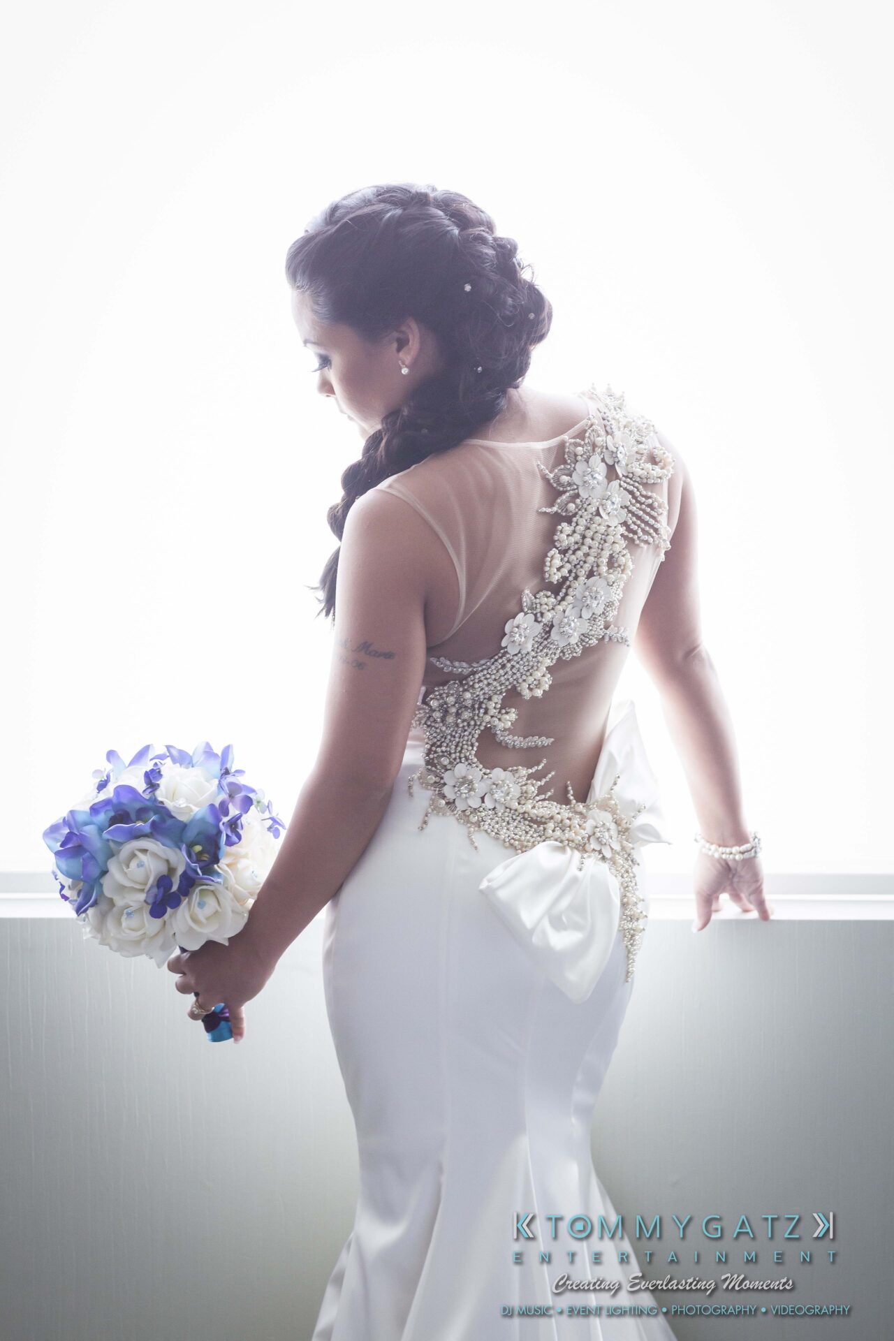 301 - TOMMY GATZ ENTERTAINMENT A bride in a white gown with intricate lace detail and a bow holds a bouquet of blue and white flowers, standing near a bright window.