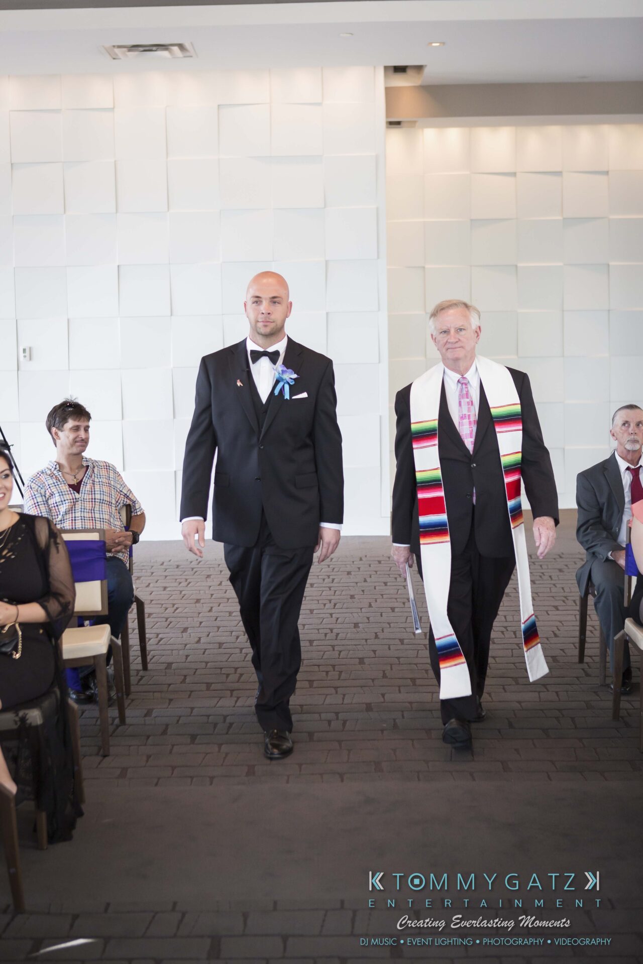 351 - TOMMY GATZ ENTERTAINMENT A groom and an officiant walk down a carpeted aisle in a brightly lit venue with seated guests on either side.