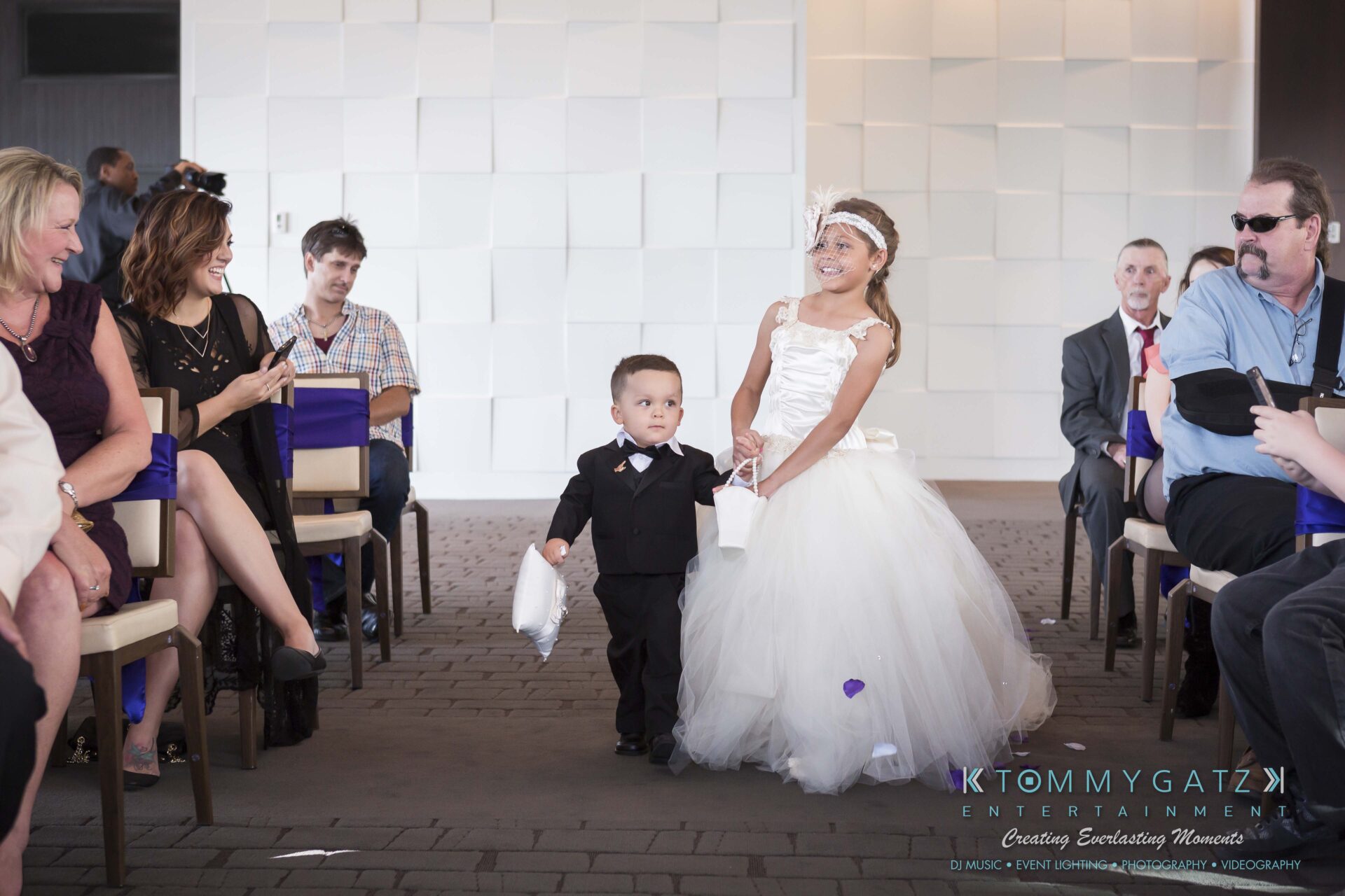 371 - TOMMY GATZ ENTERTAINMENT A young boy in a suit and a girl in a white dress walk down an aisle, surrounded by seated guests.