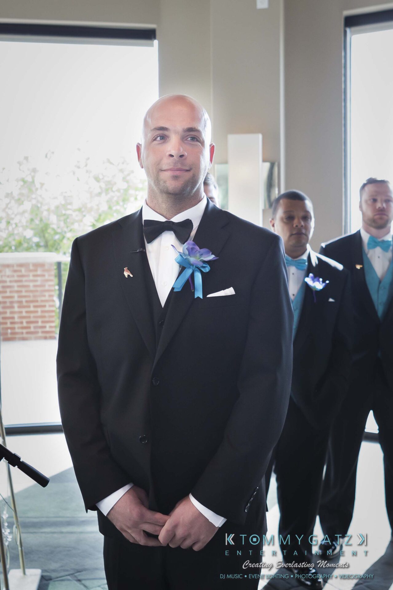 381 - TOMMY GATZ ENTERTAINMENT A groom in a tuxedo stands in a brightly lit room, wearing a blue boutonniere. Groomsmen in suits stand in the background.
