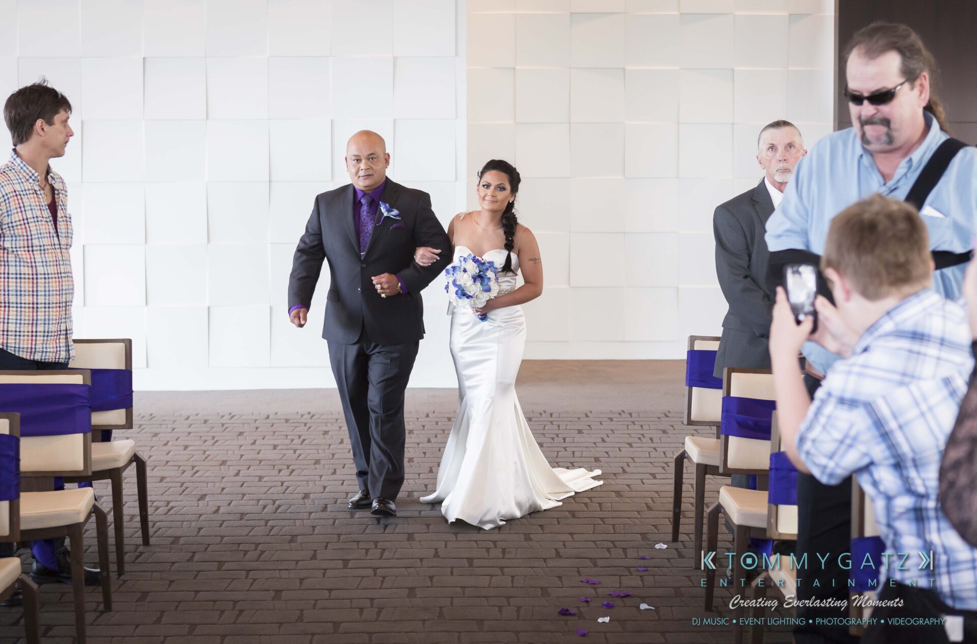 391 - TOMMY GATZ ENTERTAINMENT A bride in a white dress walks down the aisle with a man in a suit. Attendees observe from seats along the aisle.