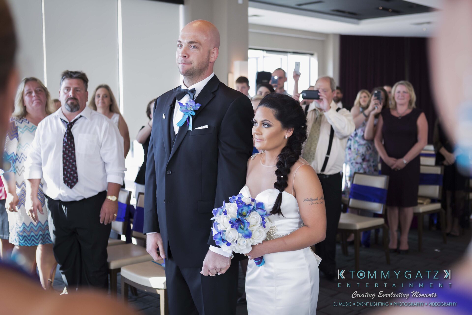 401 - TOMMY GATZ ENTERTAINMENT A bride and groom stand holding hands at their wedding ceremony, surrounded by guests.