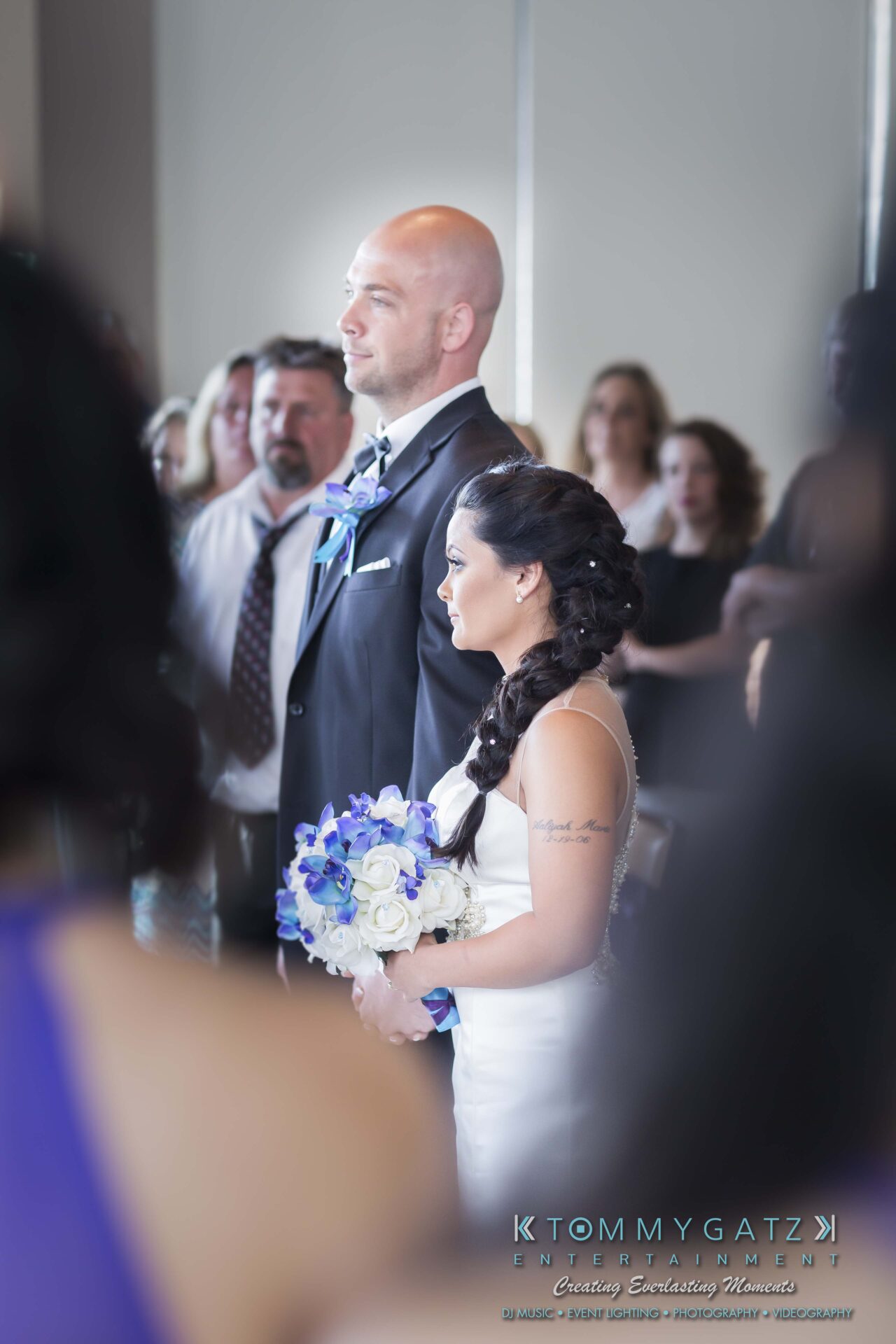 411 - TOMMY GATZ ENTERTAINMENT A bride and groom stand together indoors during a wedding ceremony, surrounded by guests. The bride holds a bouquet of white and blue flowers.