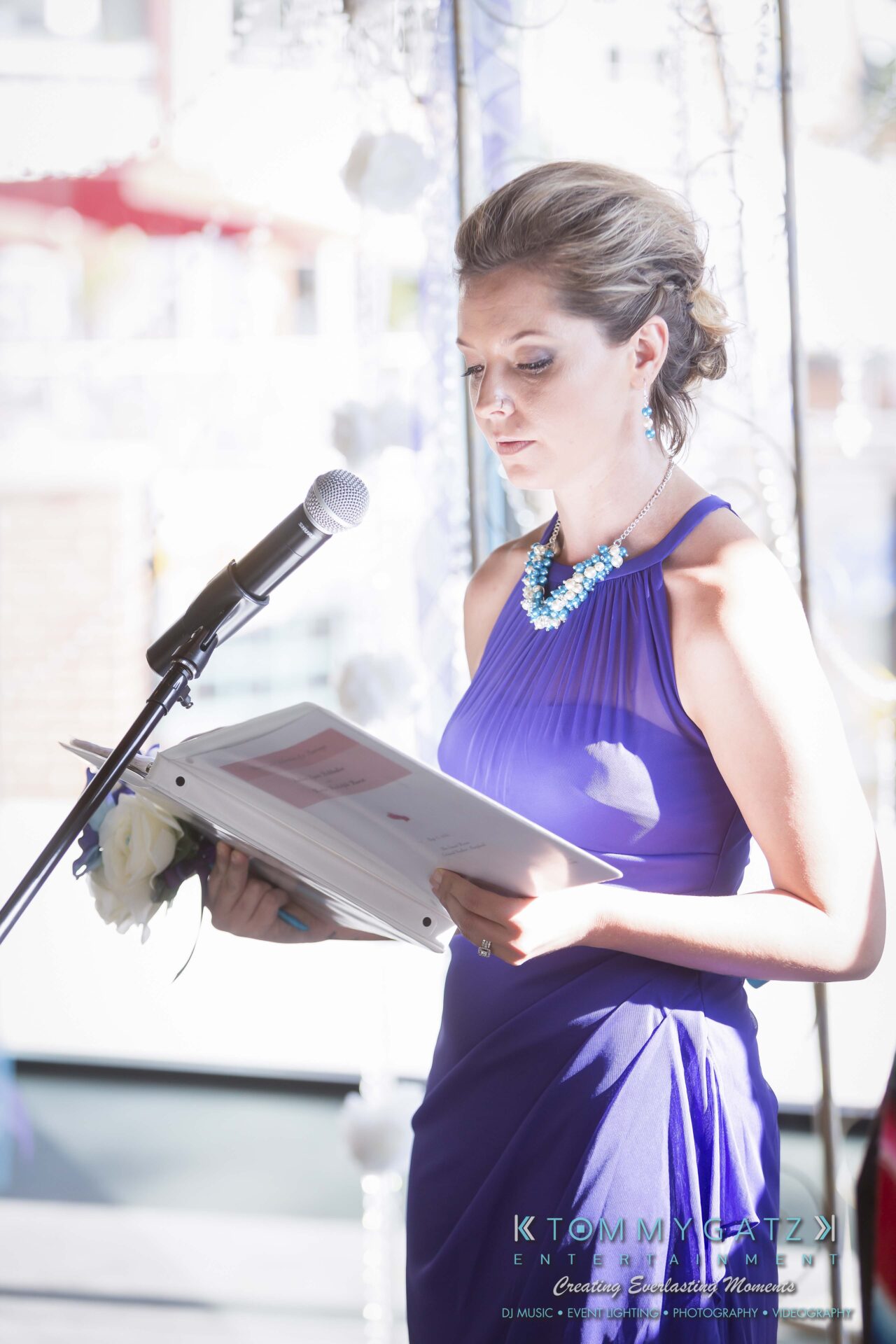 421 - TOMMY GATZ ENTERTAINMENT A woman in a blue dress stands at a microphone holding papers, possibly giving a speech, indoors with bright lighting.