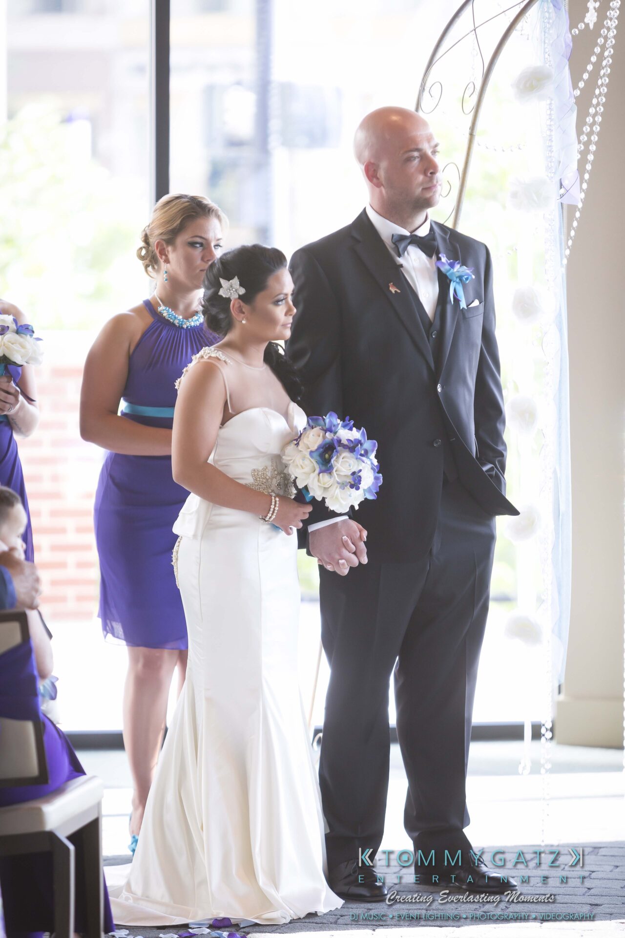 431 - TOMMY GATZ ENTERTAINMENT A bride and groom stand together, holding hands, during a wedding ceremony. The bride holds a bouquet, and bridesmaids are in the background.