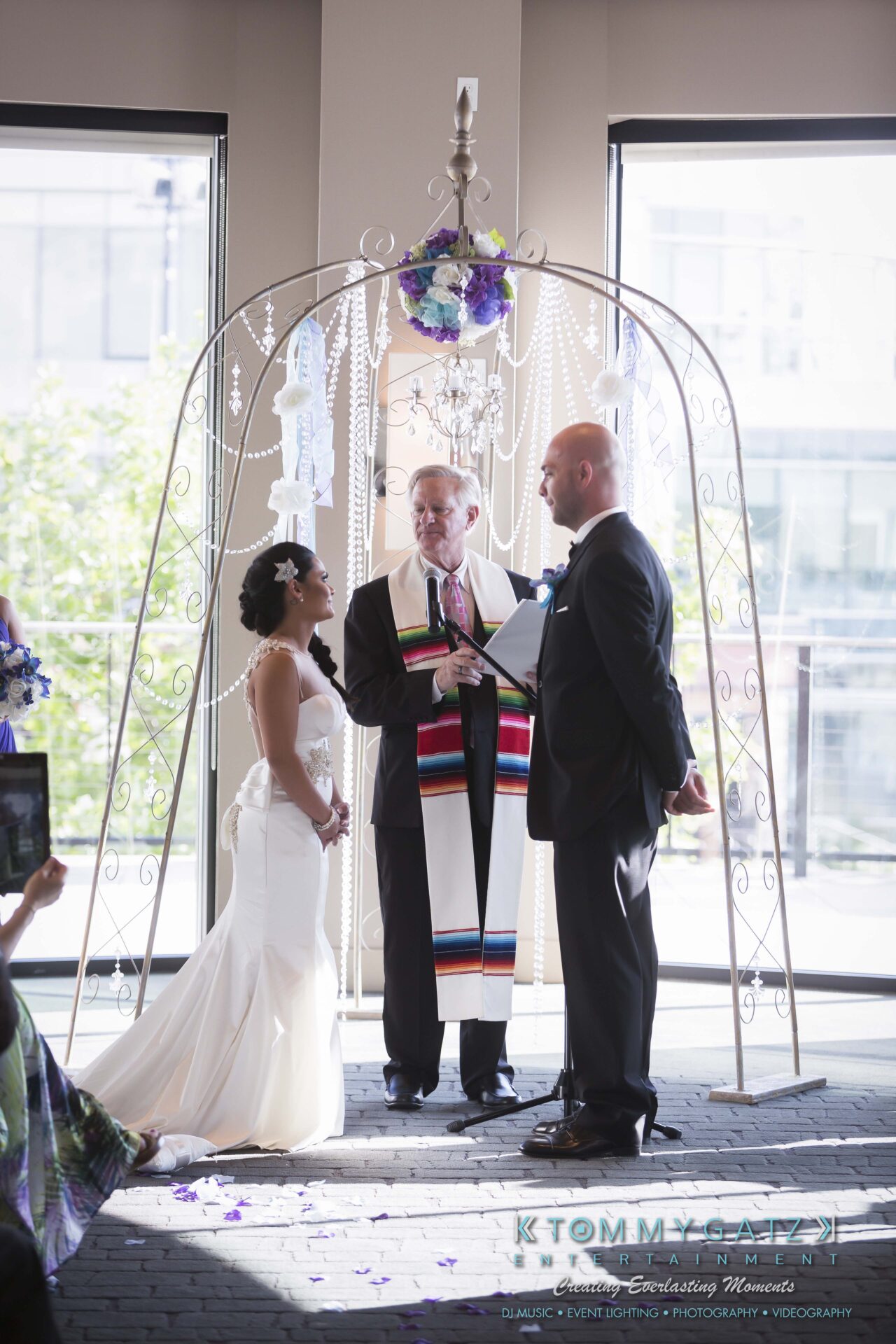 441 - TOMMY GATZ ENTERTAINMENT A couple stands under an arch, smiling at each other, while an officiant presides over their wedding ceremony indoors.