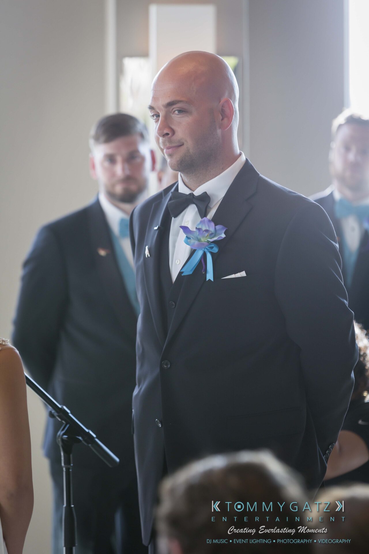 451 - TOMMY GATZ ENTERTAINMENT A man in a black suit stands smiling, wearing a boutonniere, with other people in the background at an indoor event.