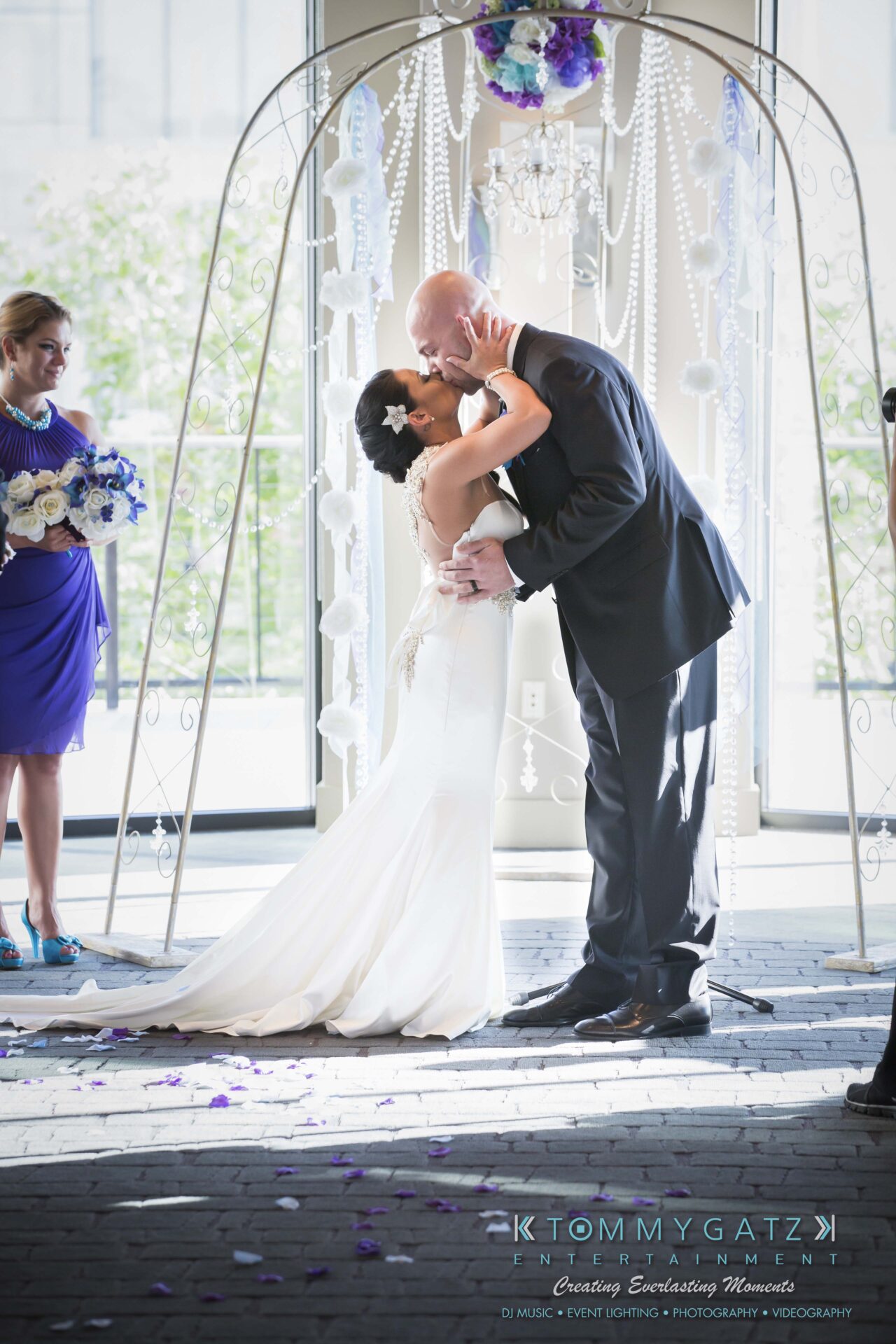 471 - TOMMY GATZ ENTERTAINMENT A couple kisses in wedding attire under an arch, with a woman in a blue dress holding flowers in the background.