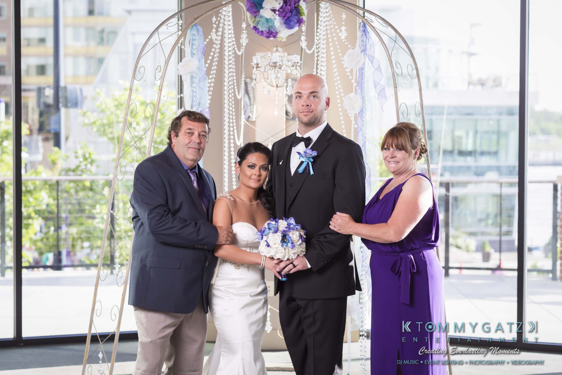 491 - TOMMY GATZ ENTERTAINMENT Four people pose under a decorative arch. The woman in a white dress holds flowers. Two others stand on either side, arm in arm. Urban setting visible through large windows in the background.