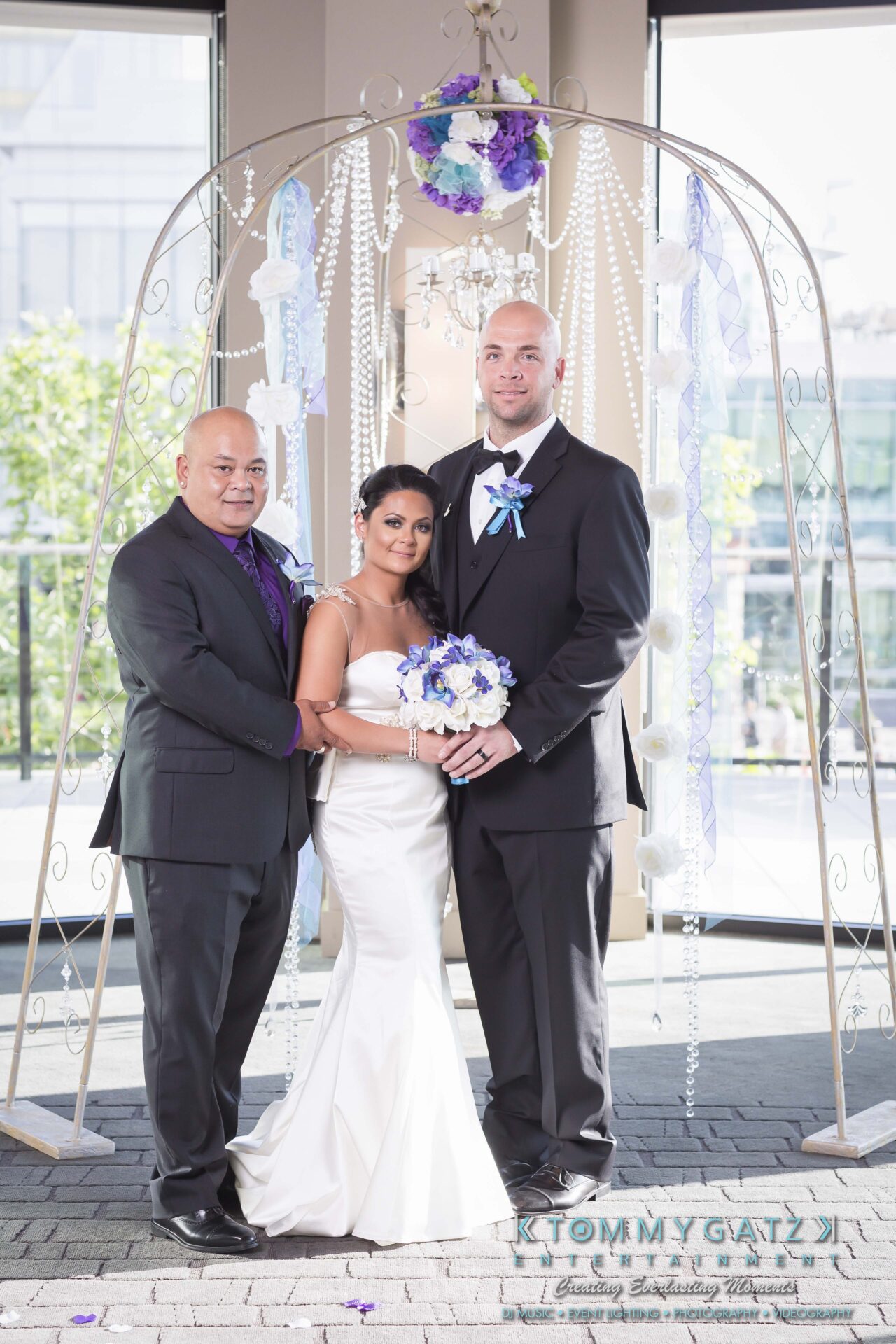 501 - TOMMY GATZ ENTERTAINMENT Three people standing under a decorated wedding arch. The woman in a white gown holds flowers, flanked by two men in suits.