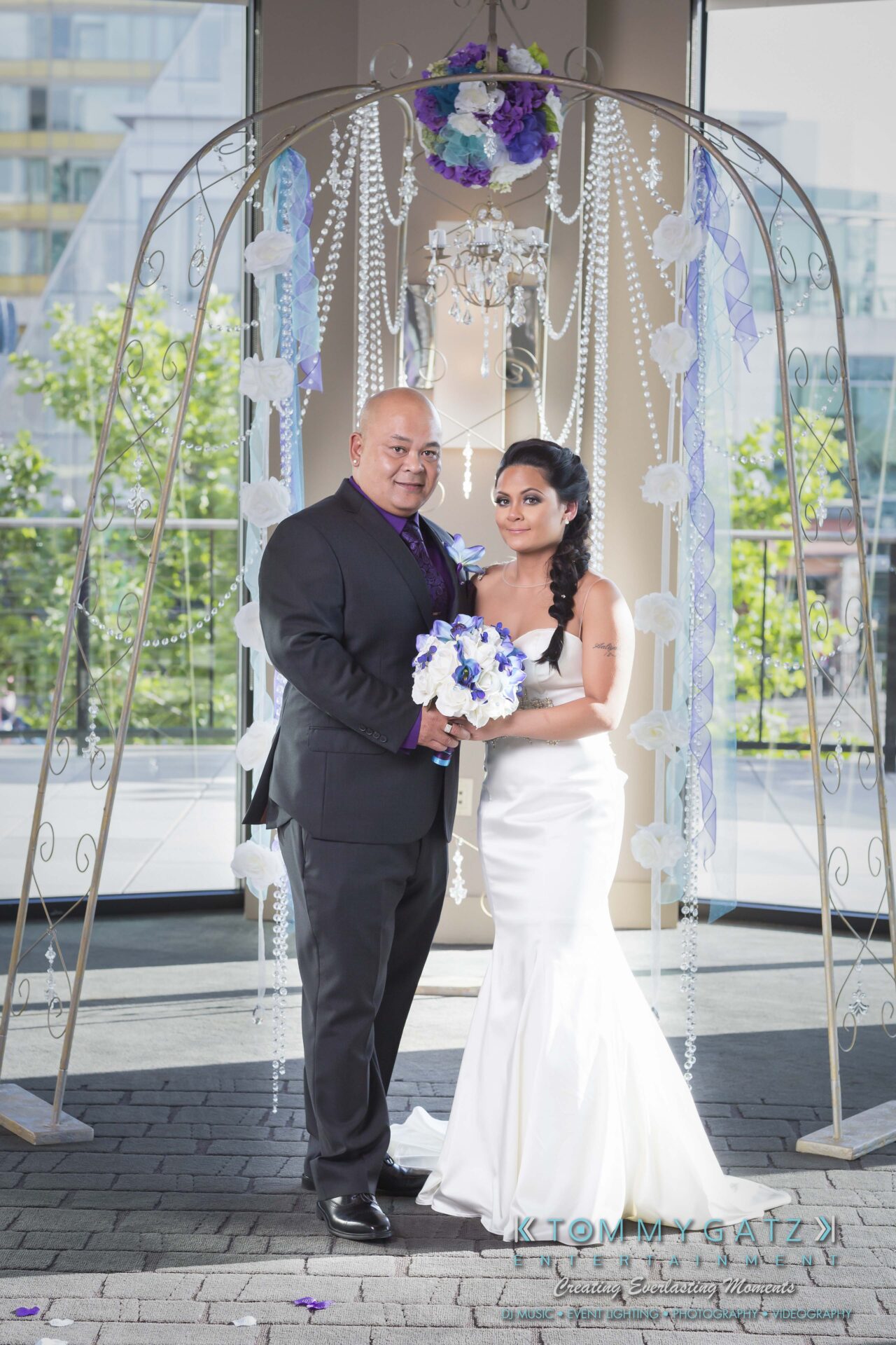 511 - TOMMY GATZ ENTERTAINMENT A couple stands under a decorative arch indoors. The woman wears a white dress and holds a bouquet, while the man wears a dark suit.