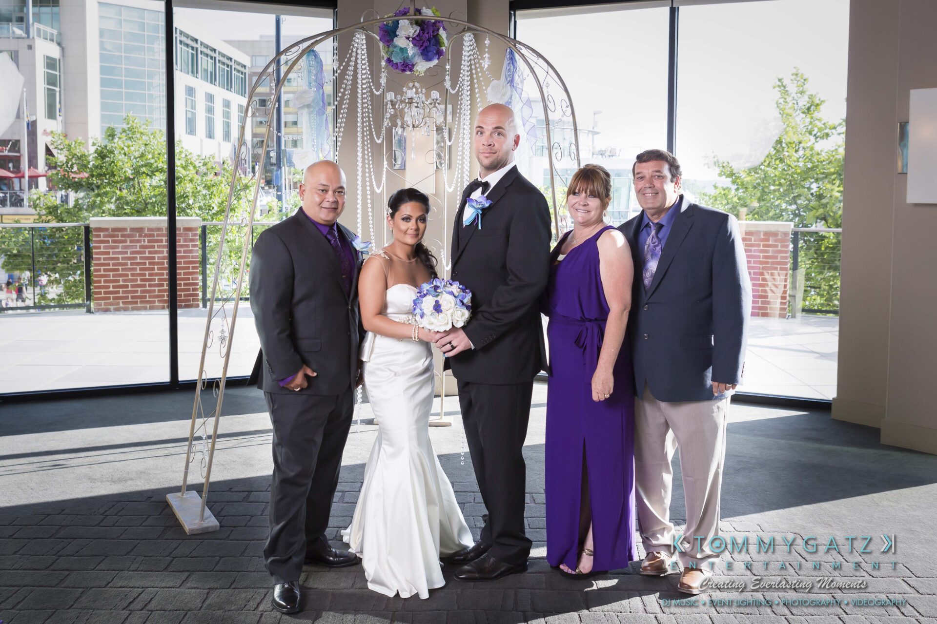 521 - TOMMY GATZ ENTERTAINMENT A bride and groom pose with three guests in a decorated indoor setting near large windows. The bride holds a bouquet of purple flowers.