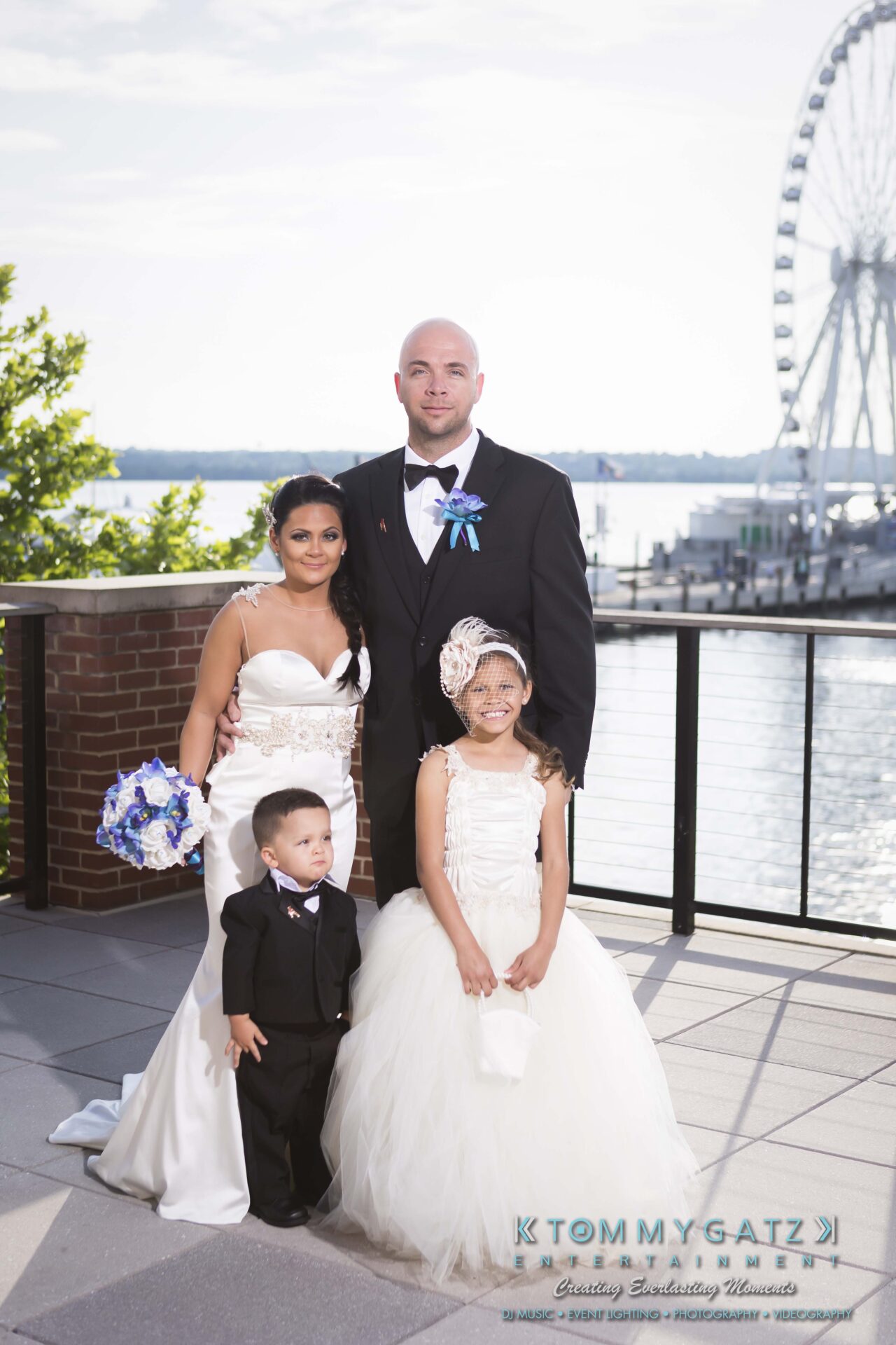 551 - TOMMY GATZ ENTERTAINMENT A wedding couple poses with two children beside a waterfront view. The girl is in a white dress, and the boy in a suit. A Ferris wheel and bridge are visible in the background.