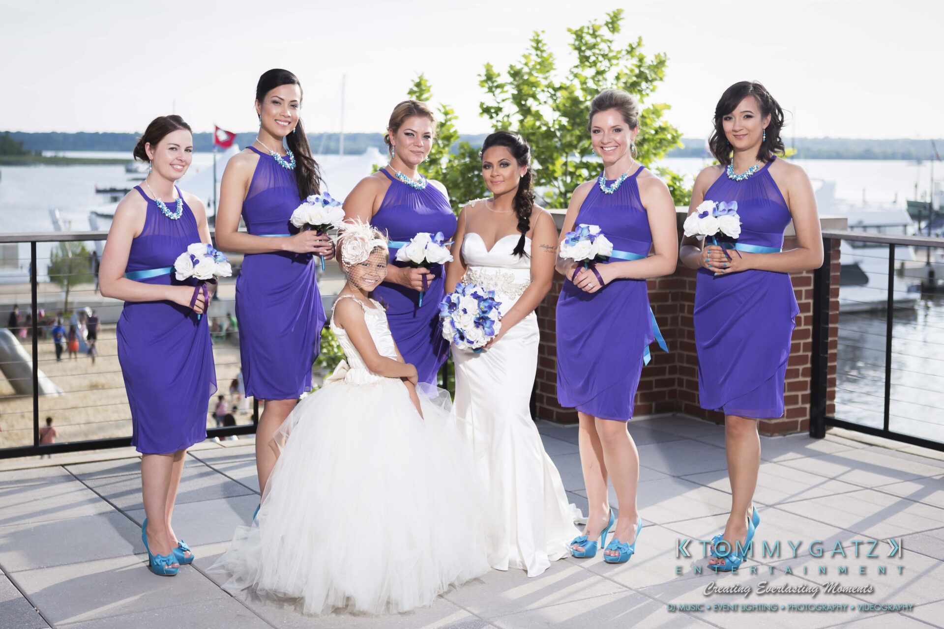 561 - TOMMY GATZ ENTERTAINMENT Bride in a white dress poses with five bridesmaids in purple dresses and a flower girl in a white dress on a waterfront terrace.