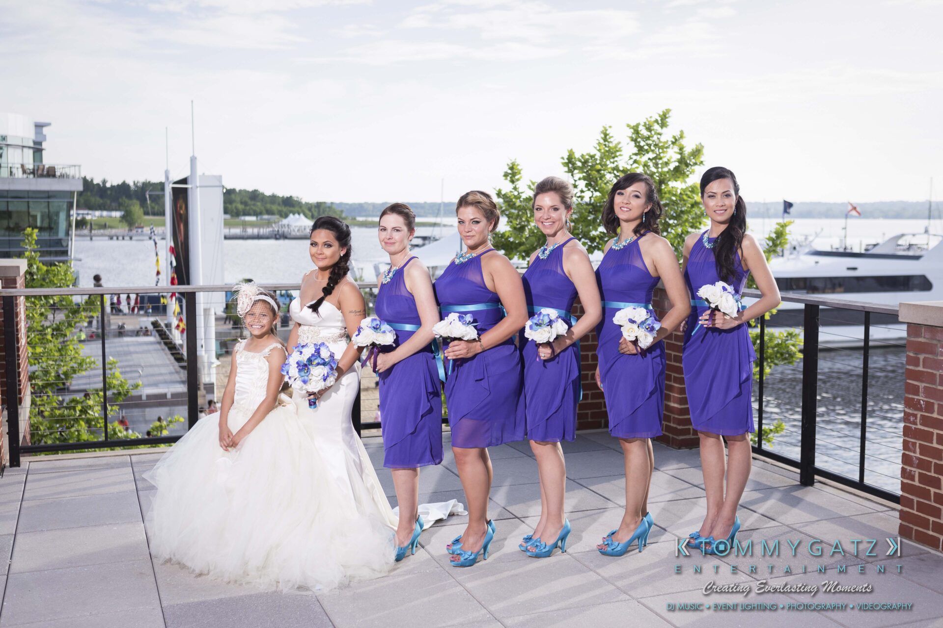 571 - TOMMY GATZ ENTERTAINMENT A bride and five bridesmaids in purple dresses pose on a waterfront terrace, with a flower girl in a white dress standing beside them.