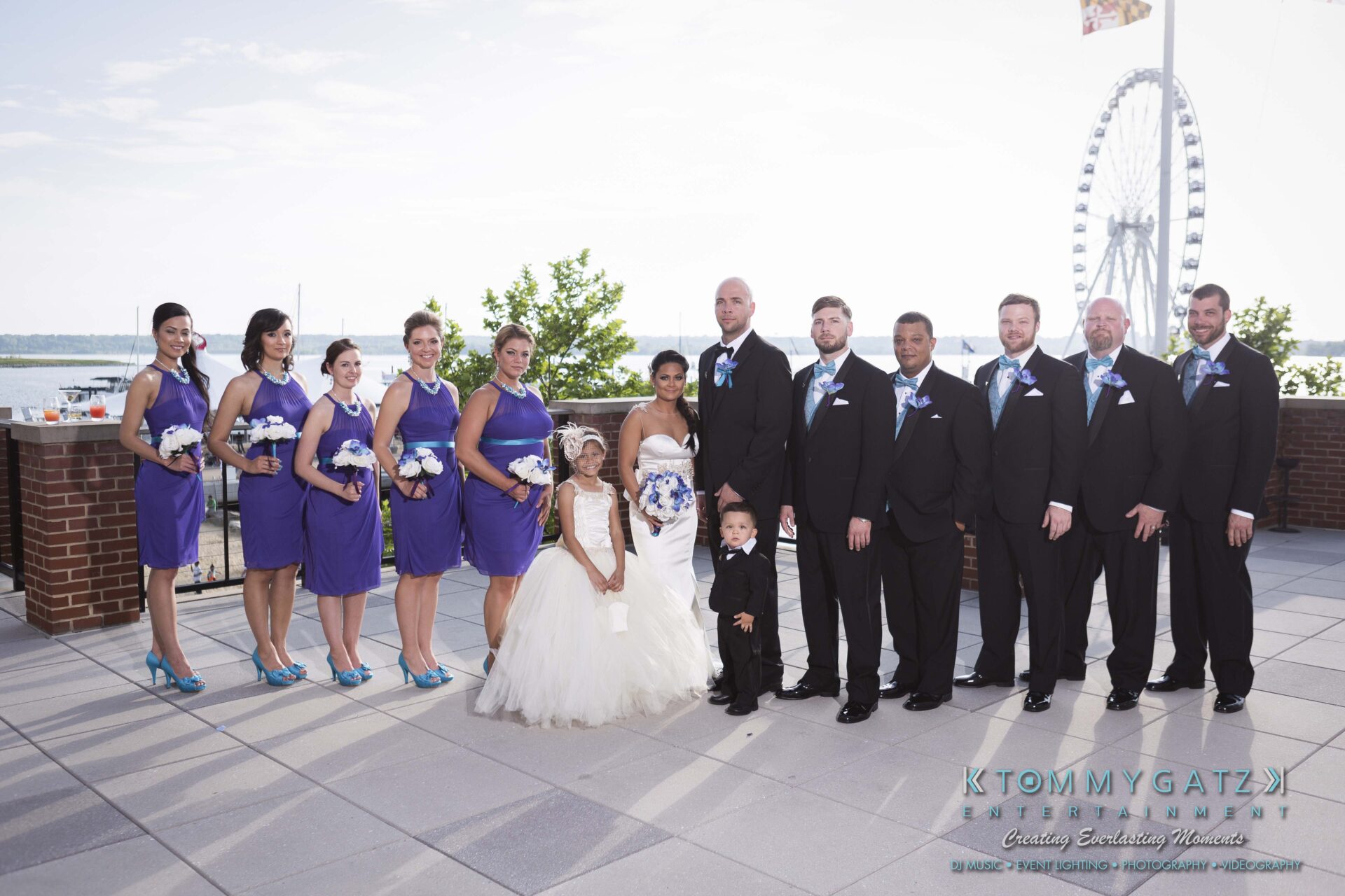 581 - TOMMY GATZ ENTERTAINMENT A group photo of a wedding party with eight adults and two children, standing outdoors near a river and ferris wheel. The women wear purple dresses and hold flowers; the men wear suits with blue ties.