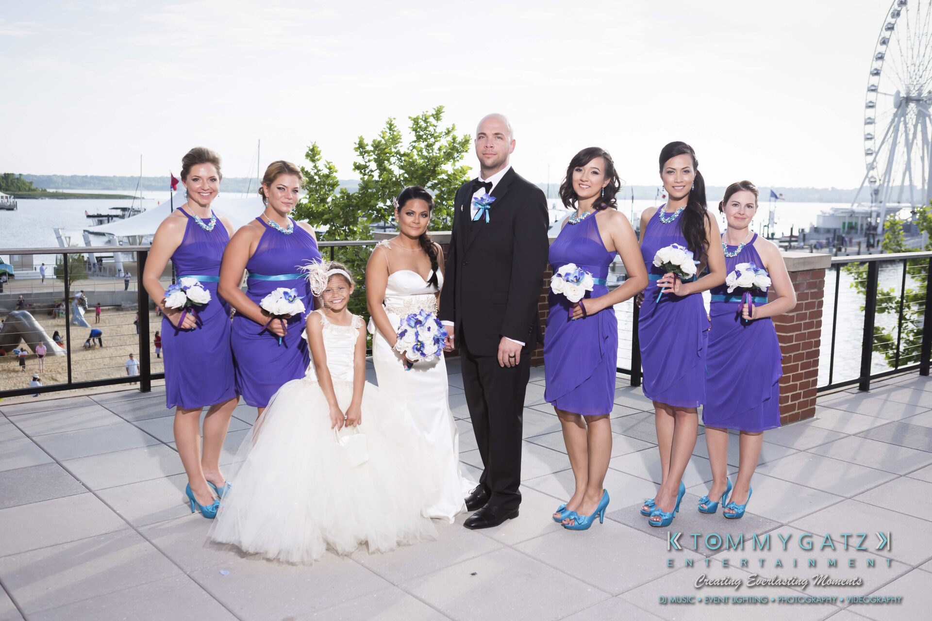 601 - TOMMY GATZ ENTERTAINMENT A bride and groom stand with a flower girl and six bridesmaids on a patio overlooking a waterfront, with a Ferris wheel in the background.
