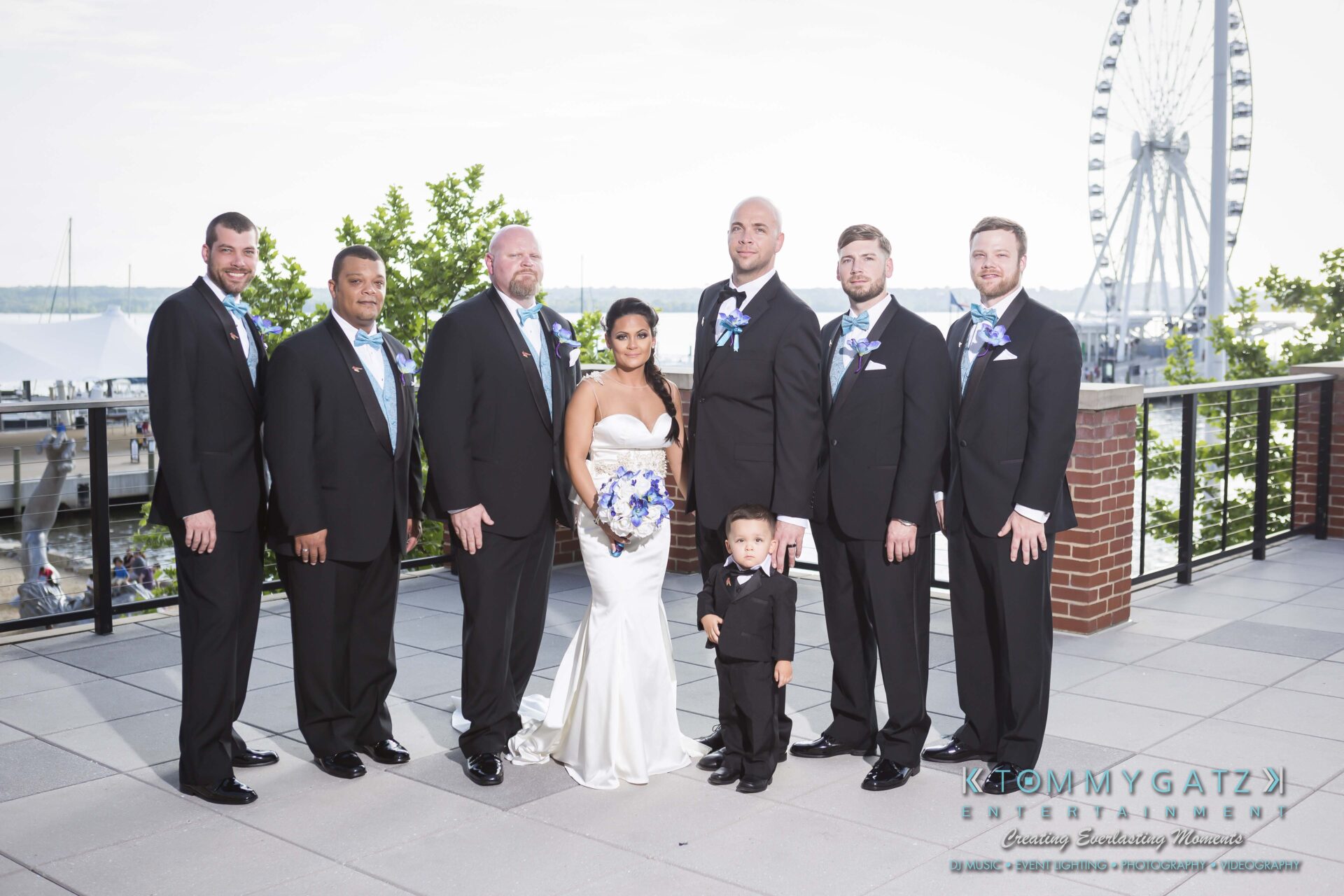 611 - TOMMY GATZ ENTERTAINMENT A bride and groom stand with five groomsmen and a child on a balcony, with a Ferris wheel and waterfront in the background.