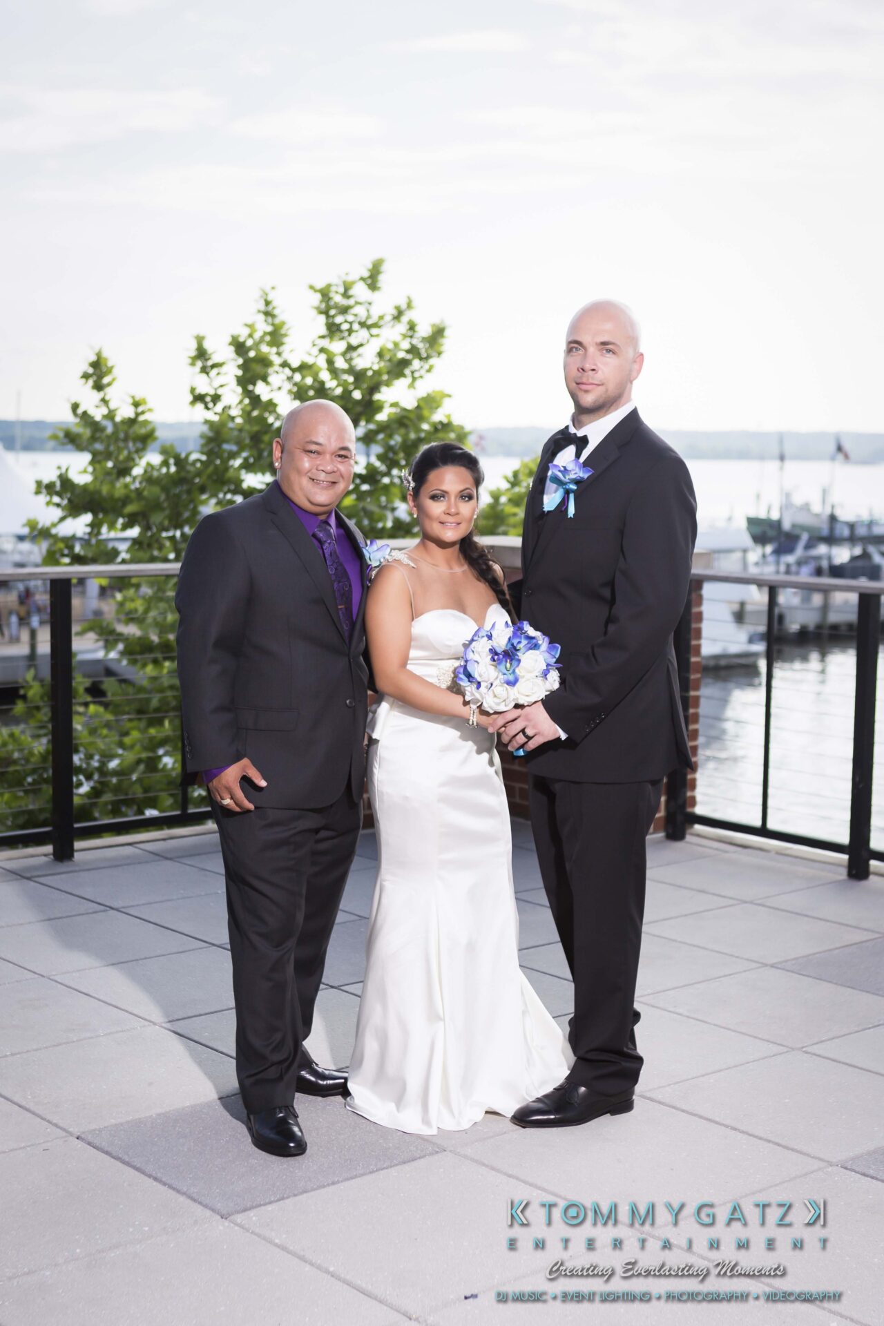641 - TOMMY GATZ ENTERTAINMENT Three people are posed on a terrace with a waterfront view. The woman in the center holds a bouquet. Two men are on either side, all dressed in formal attire.