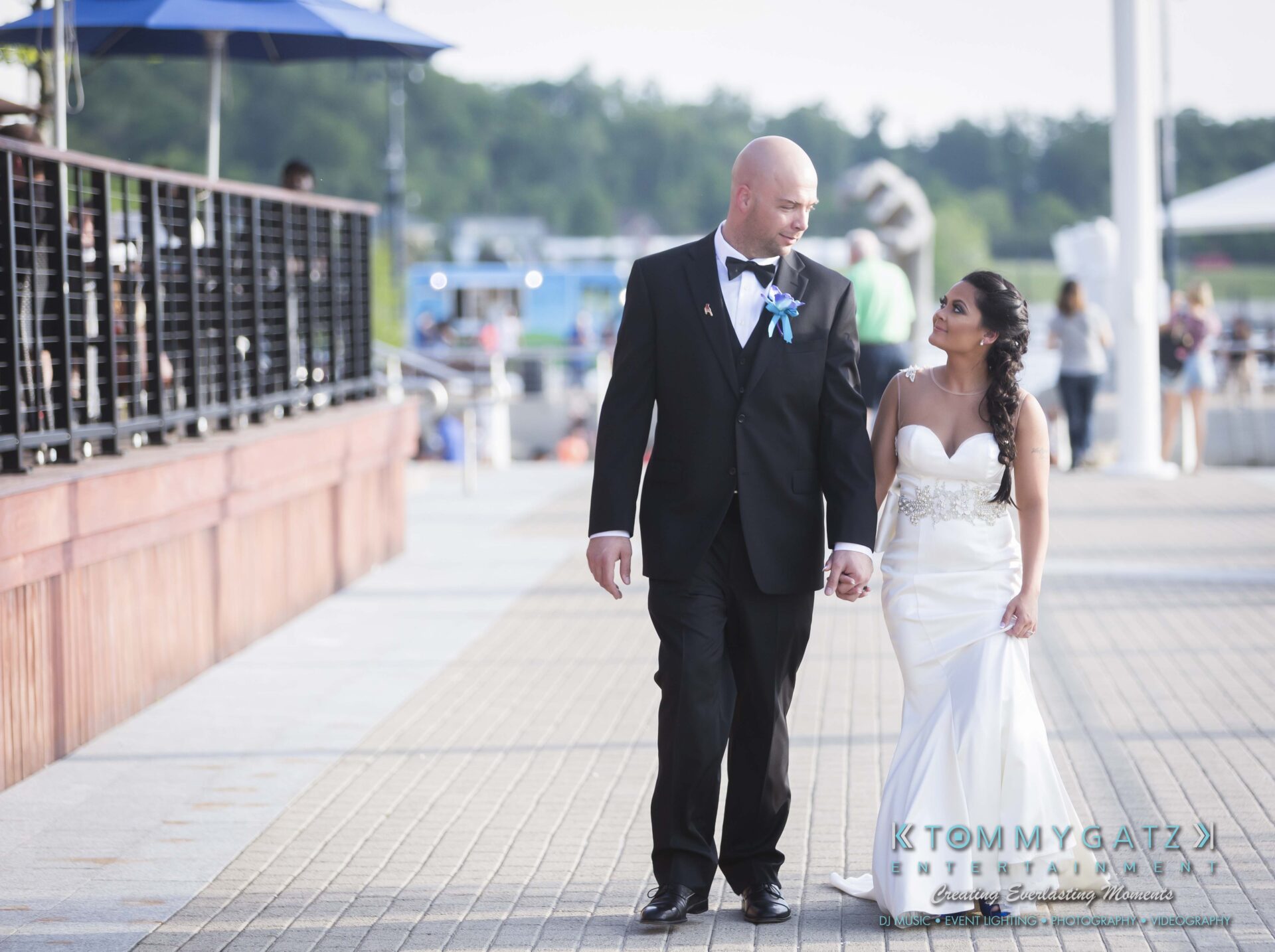 661 - TOMMY GATZ ENTERTAINMENT A bride and groom walk hand in hand along a paved path, dressed in wedding attire.
