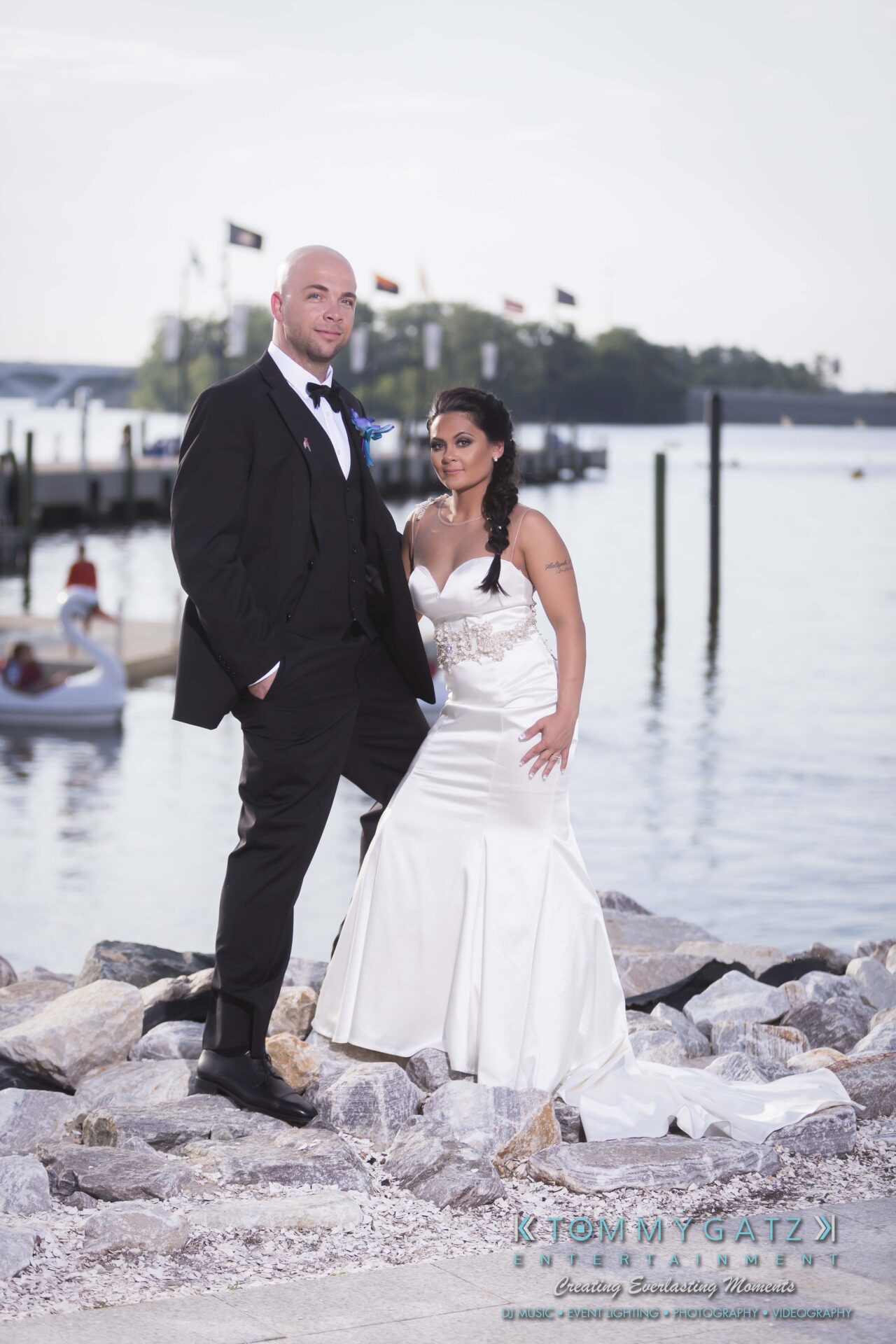 681 - TOMMY GATZ ENTERTAINMENT A couple in formal attire pose by the waterfront; the man is in a suit, and the woman wears a white gown. Flags and trees are in the background.