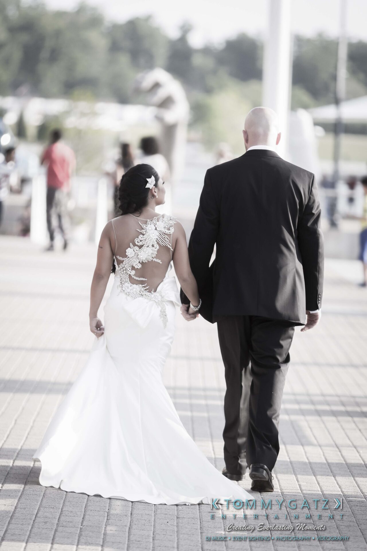 691 - TOMMY GATZ ENTERTAINMENT A couple walks hand in hand, with the woman in a white dress featuring lace details and the man in a black suit. They are outdoors on a paved pathway.