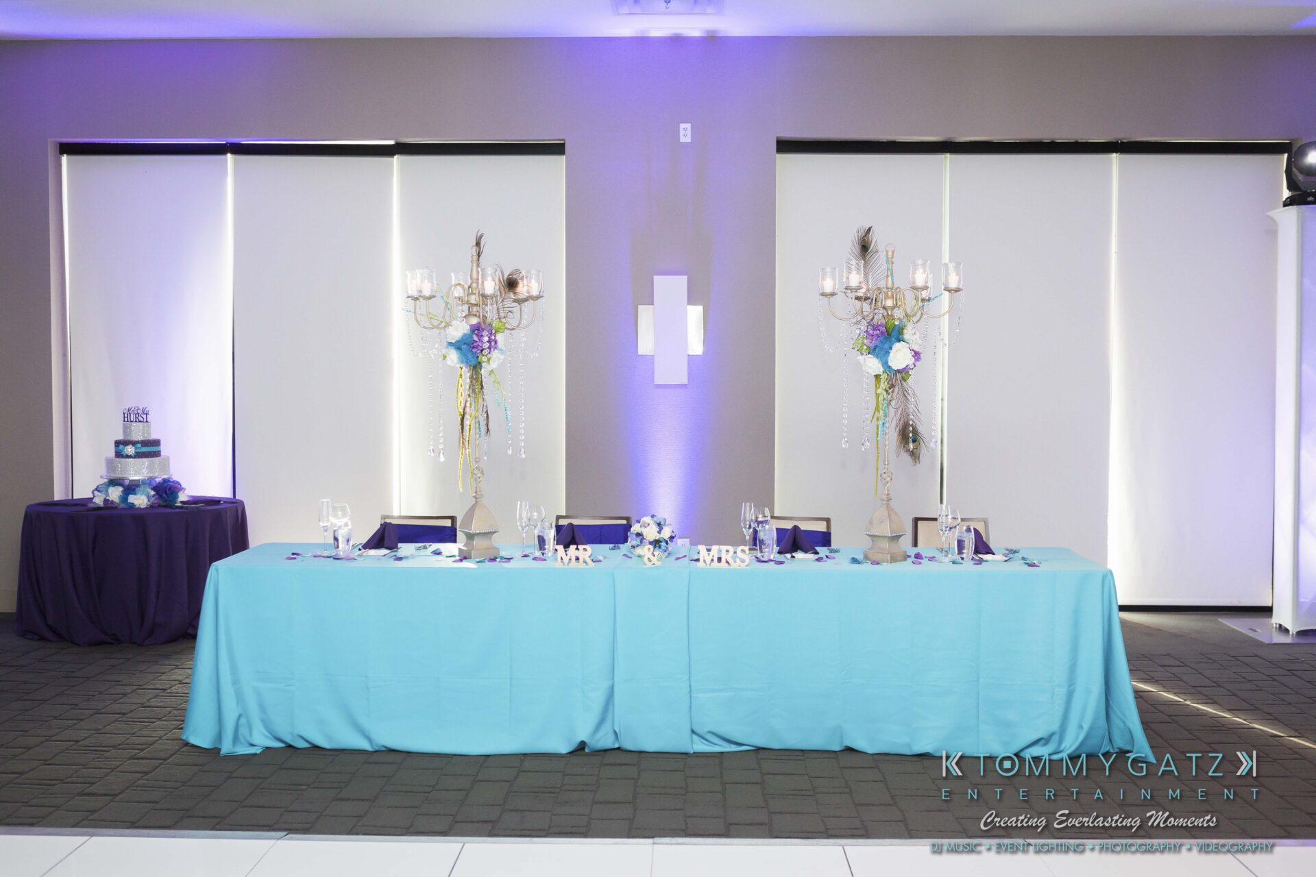701 - TOMMY GATZ ENTERTAINMENT A wedding reception table with a turquoise tablecloth, floral centerpieces, and cake table in the background. Chairs are lined up behind the table.