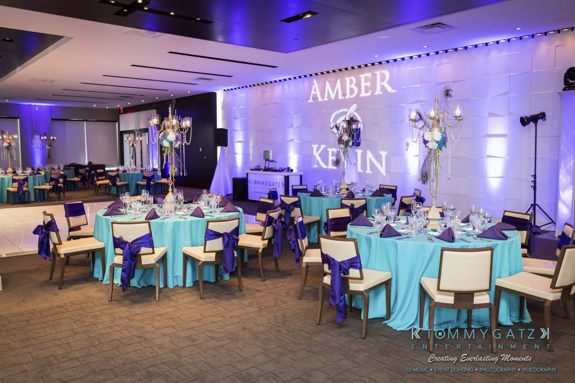 721 - TOMMY GATZ ENTERTAINMENT Elegant wedding reception setup with blue and purple decor. Tables are set with centerpiece arrangements, and a backdrop displays "Amber & Kevin.