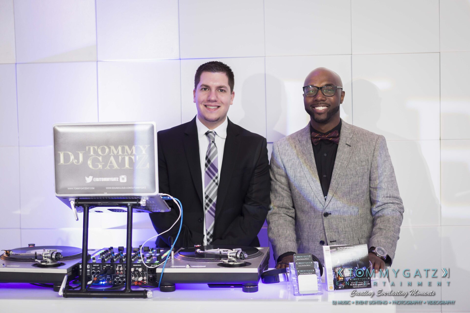 731 - TOMMY GATZ ENTERTAINMENT Two DJs standing behind a table with DJ equipment and a laptop. The table has "Tommy Gatz" branding. Both are dressed in suits and smiling.