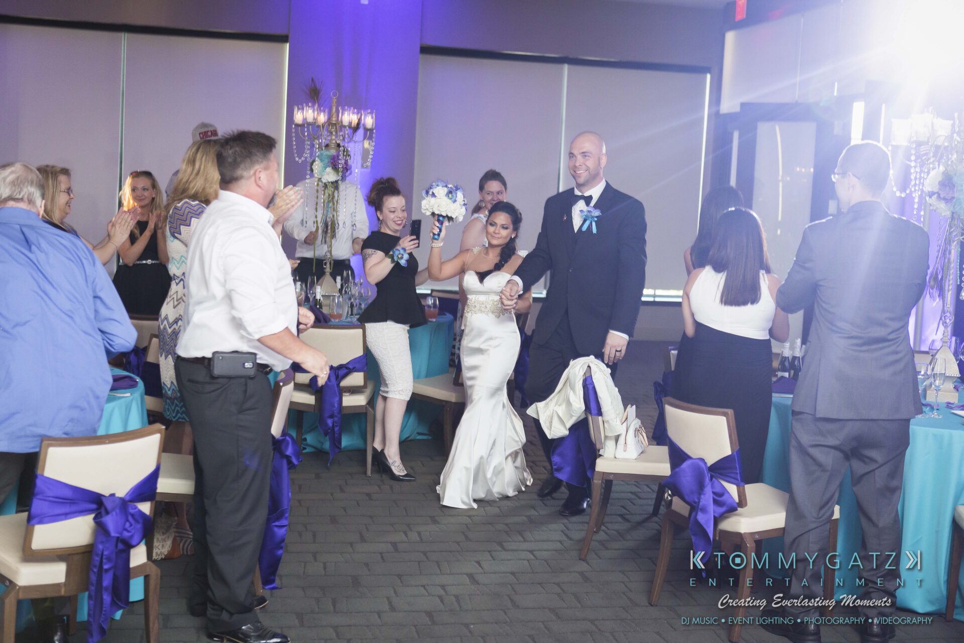 761 - TOMMY GATZ ENTERTAINMENT A bride and groom enter a reception room while guests clap and take photos.