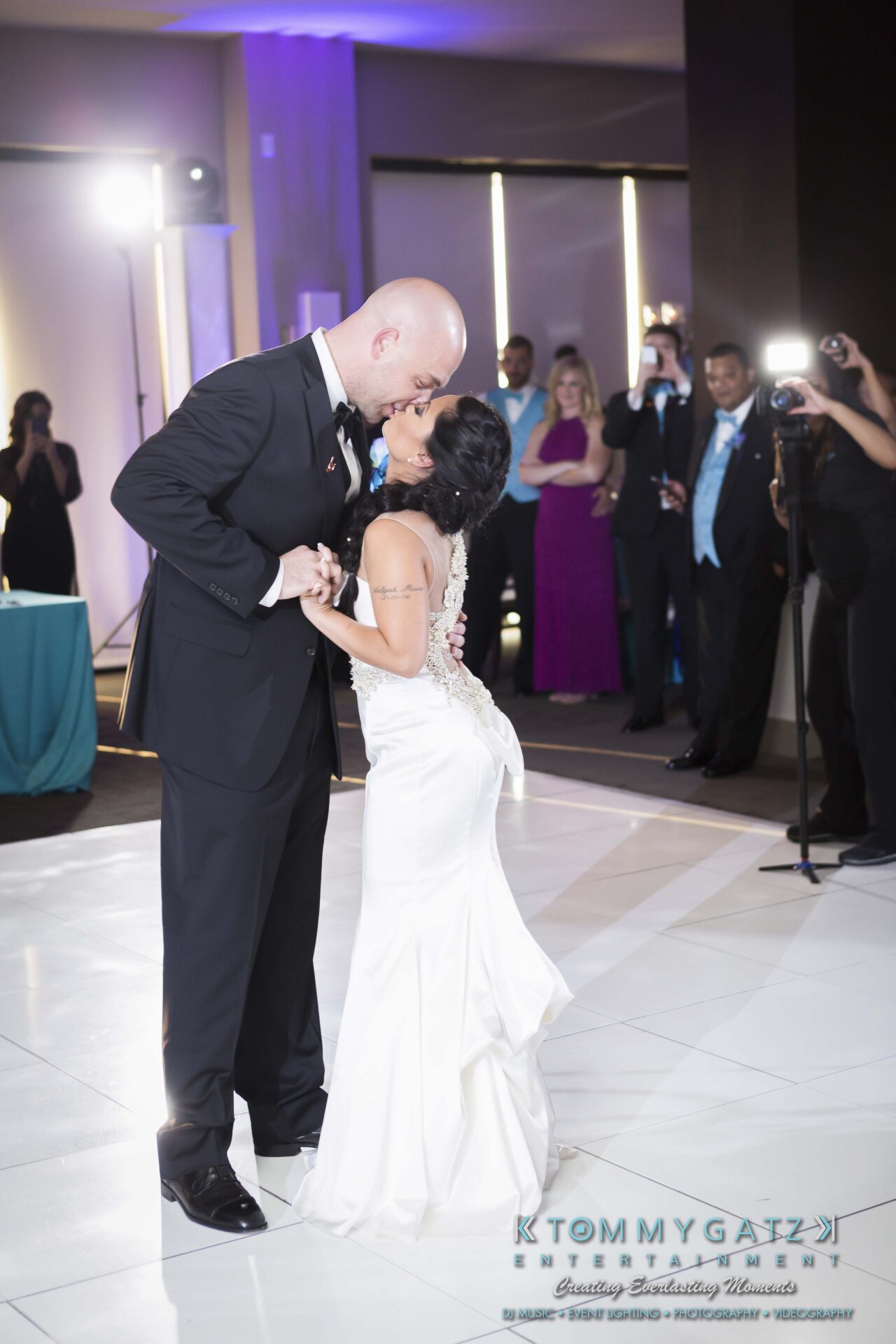 781 - TOMMY GATZ ENTERTAINMENT A couple in formal attire shares a kiss on the dance floor at a wedding reception, surrounded by guests and photographers.