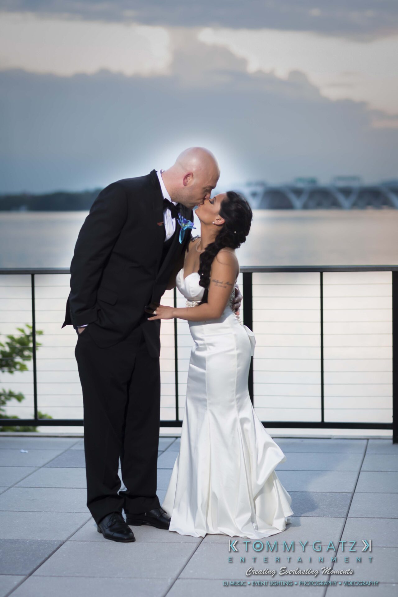 831 - TOMMY GATZ ENTERTAINMENT A couple in formal attire kisses on a terrace with a body of water and distant bridge in the background.