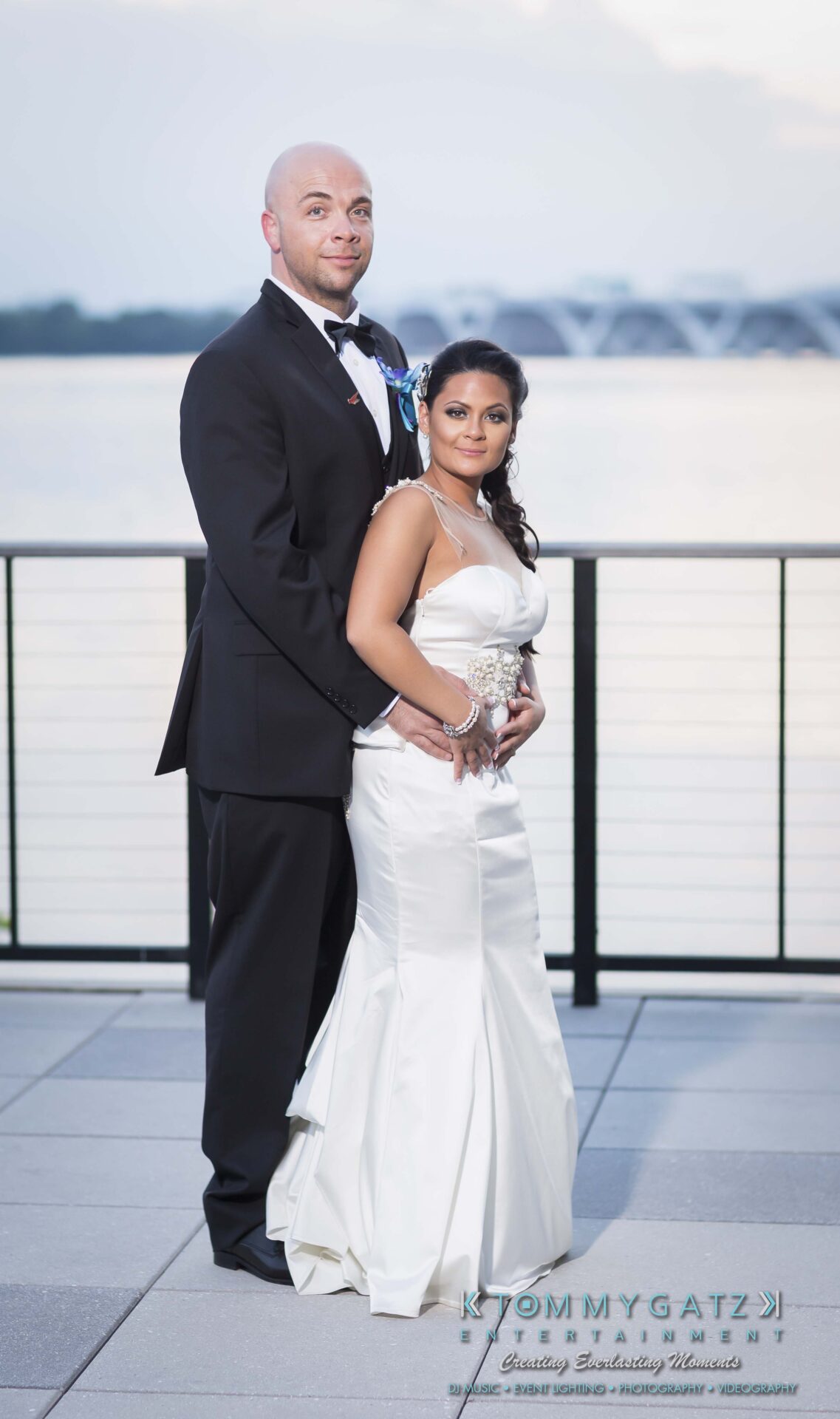 841 - TOMMY GATZ ENTERTAINMENT A couple in formal attire poses on a patio with a waterfront view. The woman is wearing a white gown, and the man is in a black suit.