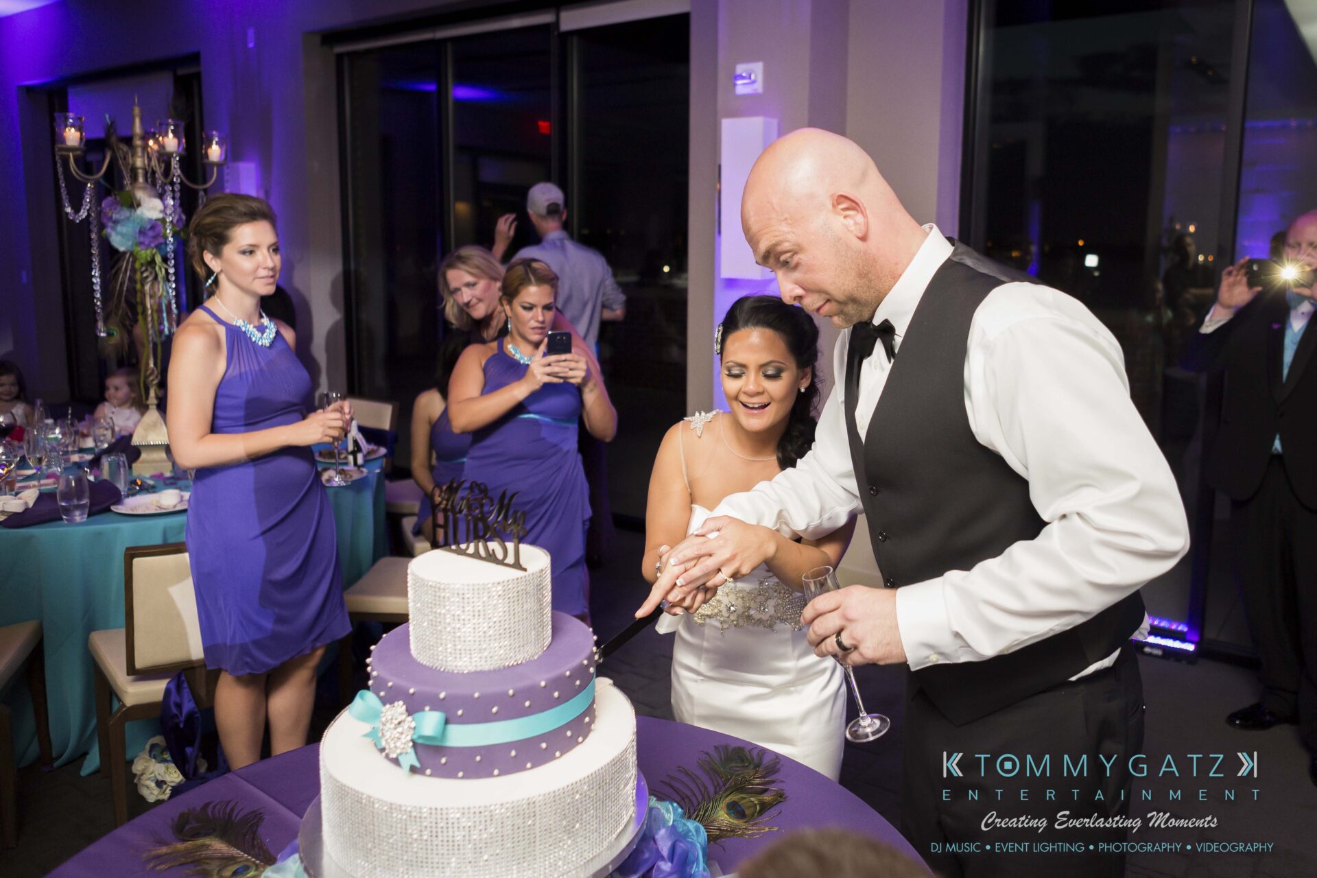 93 - TOMMY GATZ ENTERTAINMENT A groom and bride cut a tiered wedding cake at a reception. Bridesmaids in purple dresses watch, and guests take photos.