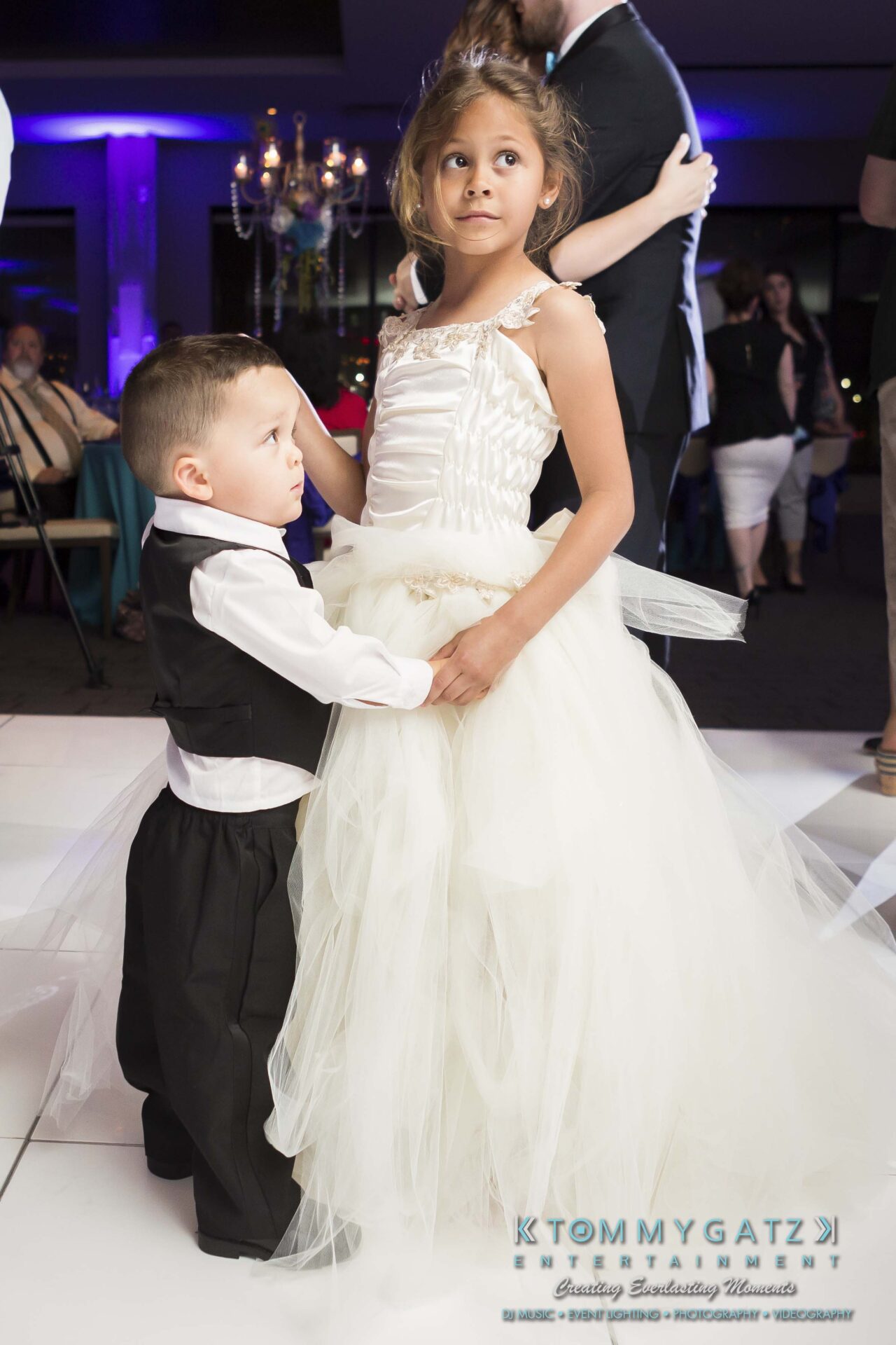 95 - TOMMY GATZ ENTERTAINMENT A young girl in a white dress and a boy in a tuxedo stand together on a dance floor during an event.