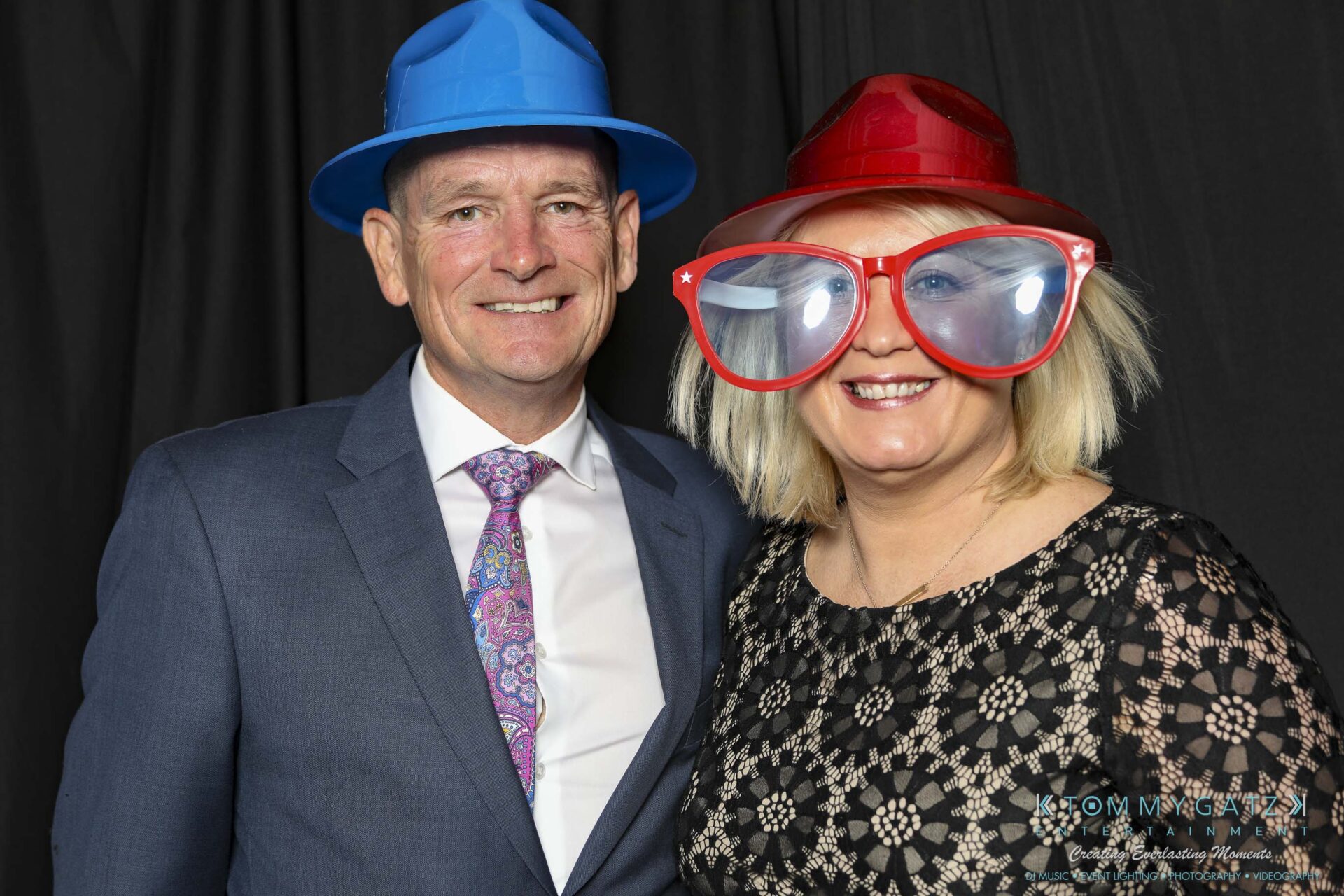 Megan-Matt-102718-Blow-WM-1 - TOMMY GATZ ENTERTAINMENT A man in a suit and a woman in a patterned dress stand smiling. The man wears a blue hat, and the woman wears a red hat and large novelty glasses against a black backdrop.