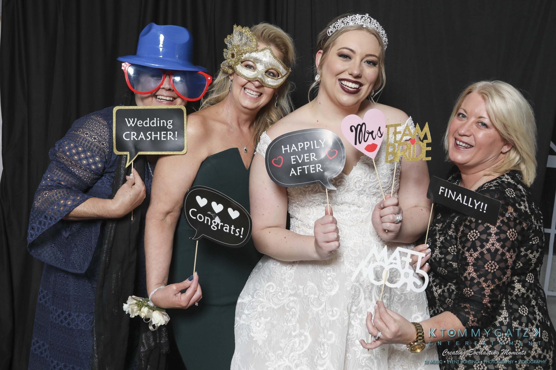 Megan-Matt-102718-Blow-WM-13 - TOMMY GATZ ENTERTAINMENT Four women posing at a photo booth. One is in a wedding dress. They're holding festive signs, wearing accessories like hats and masks. The background is black.