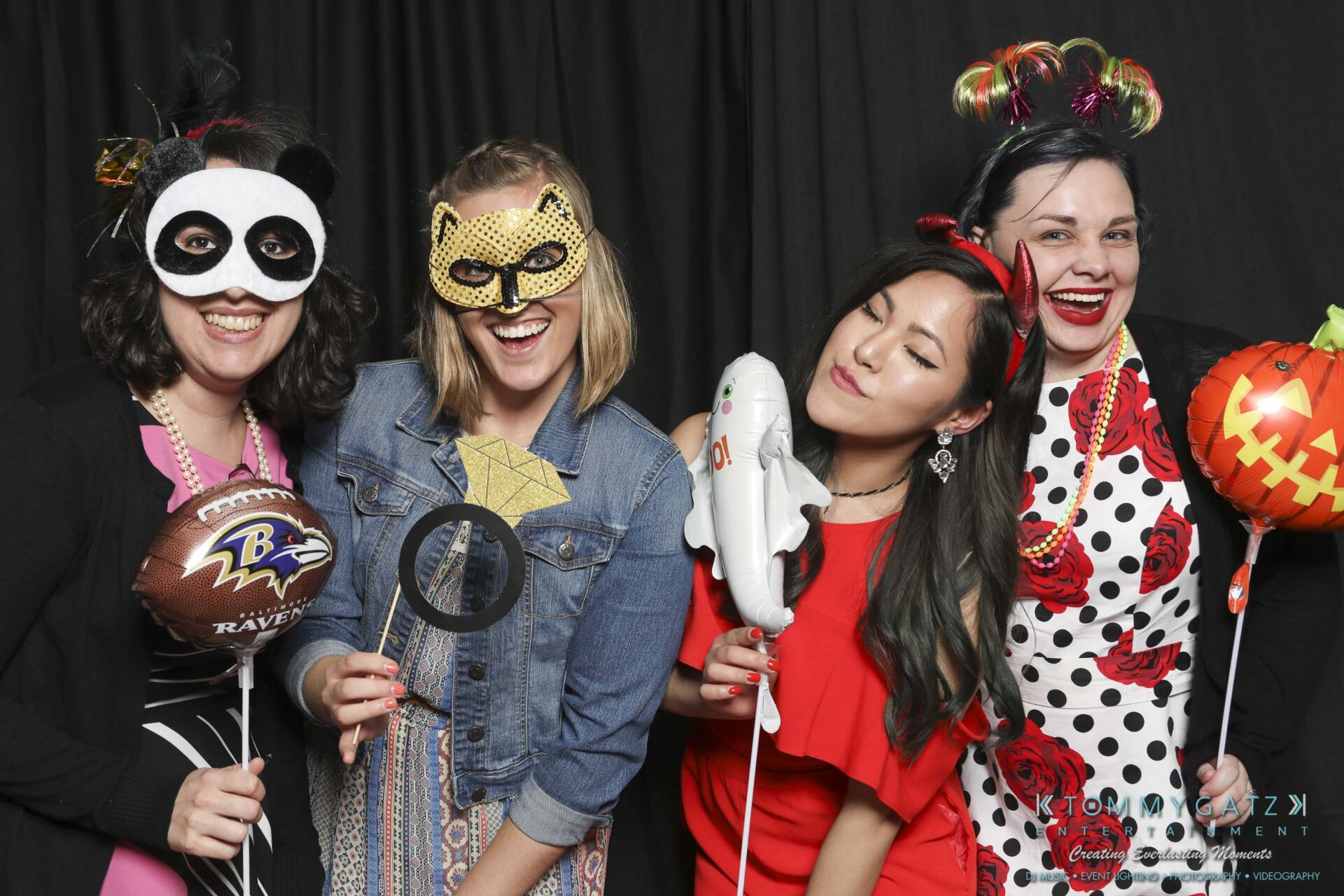 Megan-Matt-102718-Blow-WM-16 - TOMMY GATZ ENTERTAINMENT Four people in colorful costumes and masks pose in front of a black curtain holding fun props, including a football, dinosaur, and jack-o'-lantern, smiling and enjoying themselves.