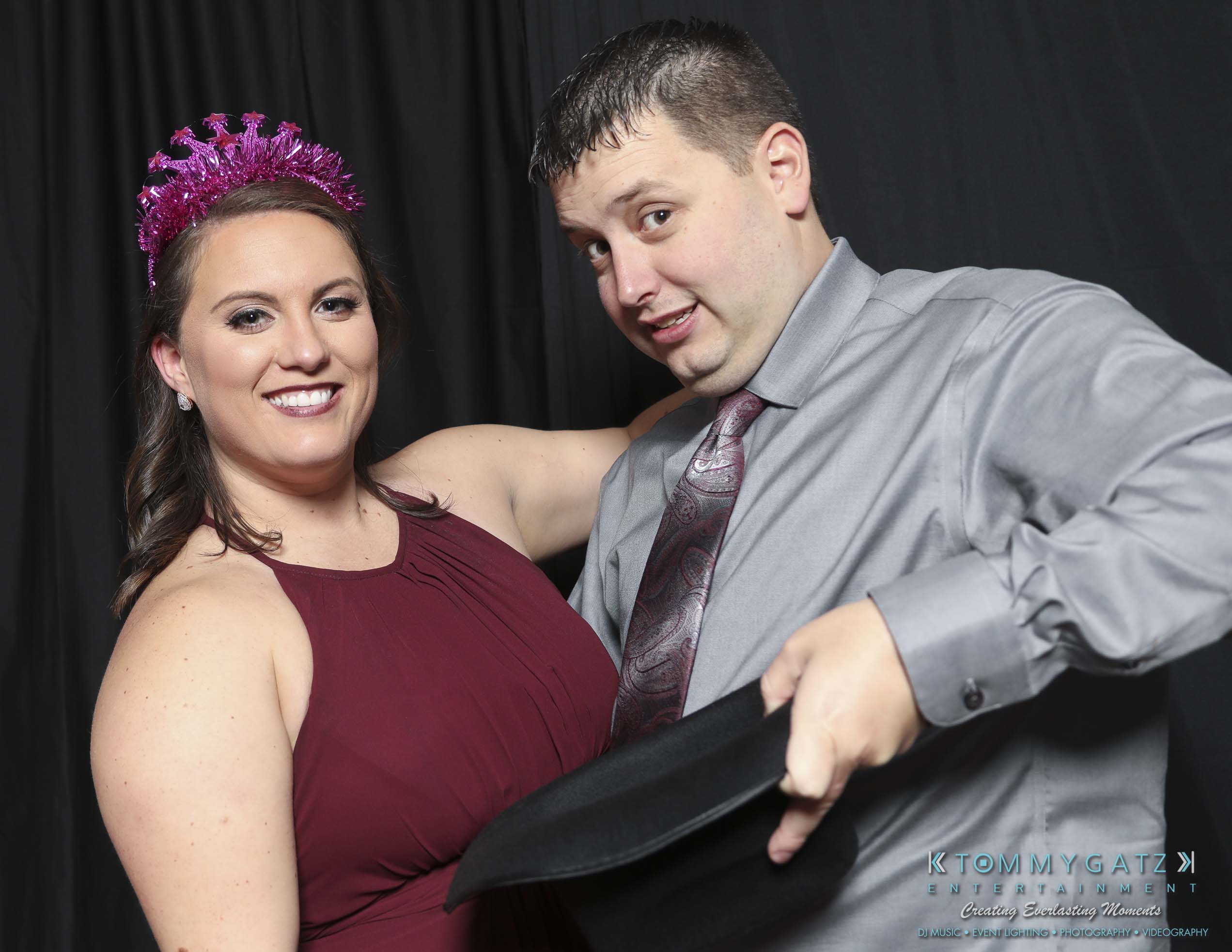 Megan-Matt-102718-Blow-WM-19 - TOMMY GATZ ENTERTAINMENT A man and woman smile at the camera; the woman wears a purple tiara and a sleeveless dress, while the man is in a gray shirt holding a black hat against a dark background.