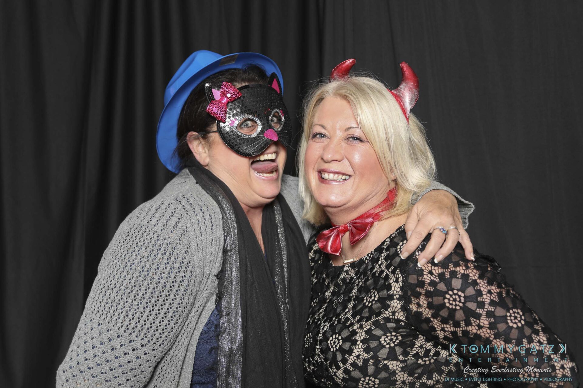 Megan-Matt-102718-Blow-WM-2 - TOMMY GATZ ENTERTAINMENT Two smiling women posing together; one wears a cat mask and blue hat, the other has devil horns. They stand in front of a black curtain.