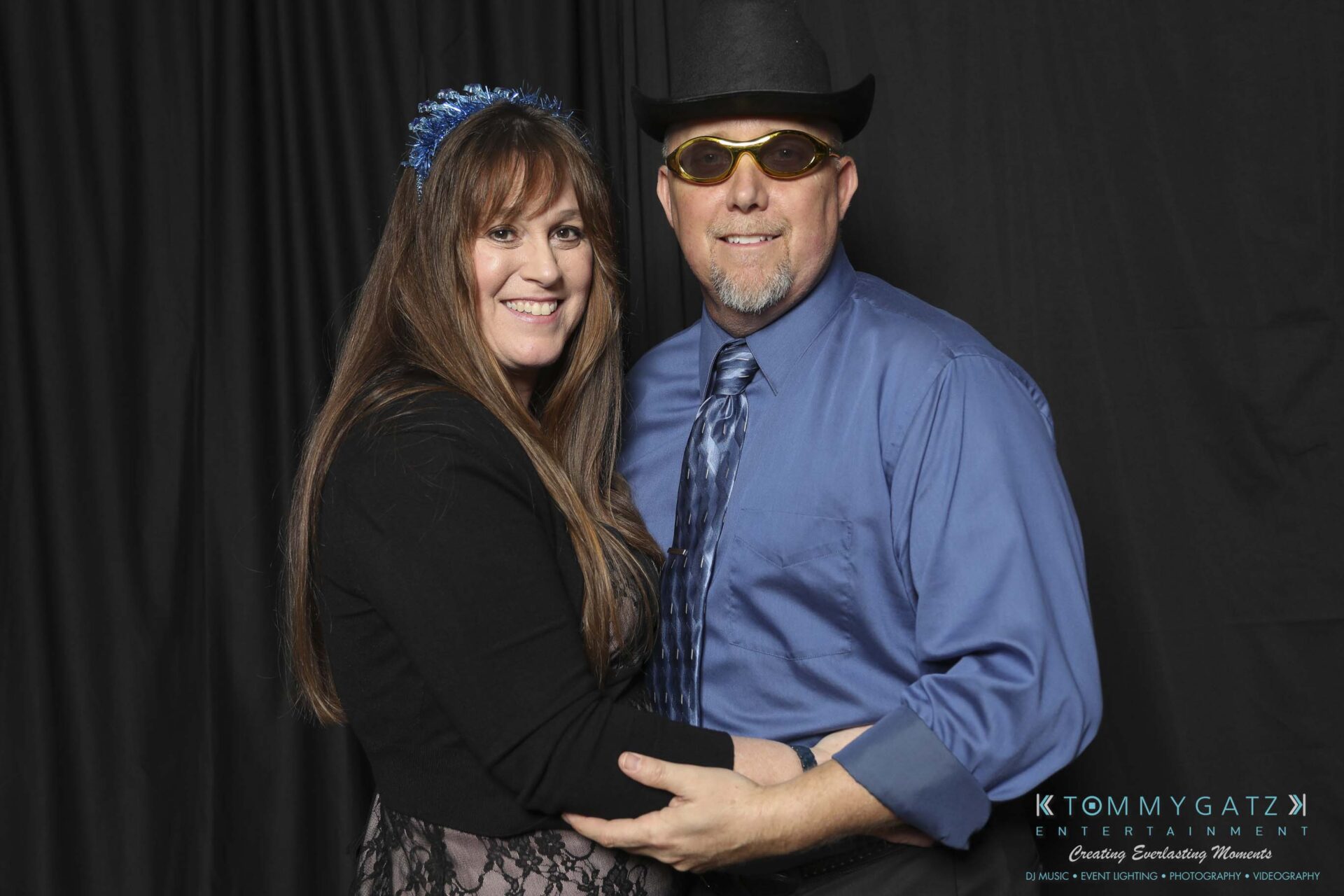 Megan-Matt-102718-Blow-WM-21 - TOMMY GATZ ENTERTAINMENT A man and woman pose together. The man wears a hat and sunglasses, while the woman wears a headband with blue accents. They stand in front of a black backdrop.