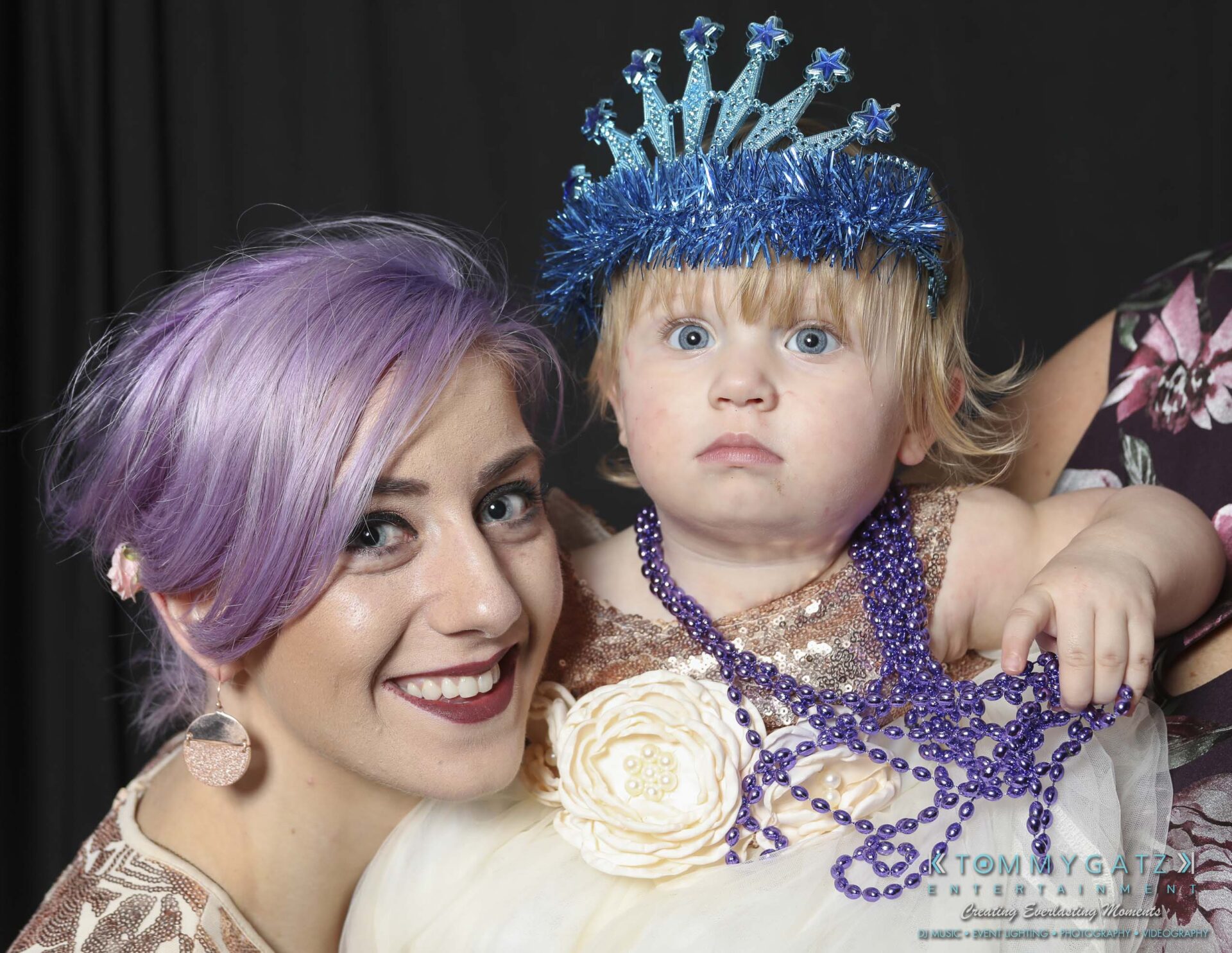 Megan-Matt-102718-Blow-WM-23 - TOMMY GATZ ENTERTAINMENT Woman with purple hair smiling, holding a child wearing a blue tinsel crown and bead necklaces, against a dark background.